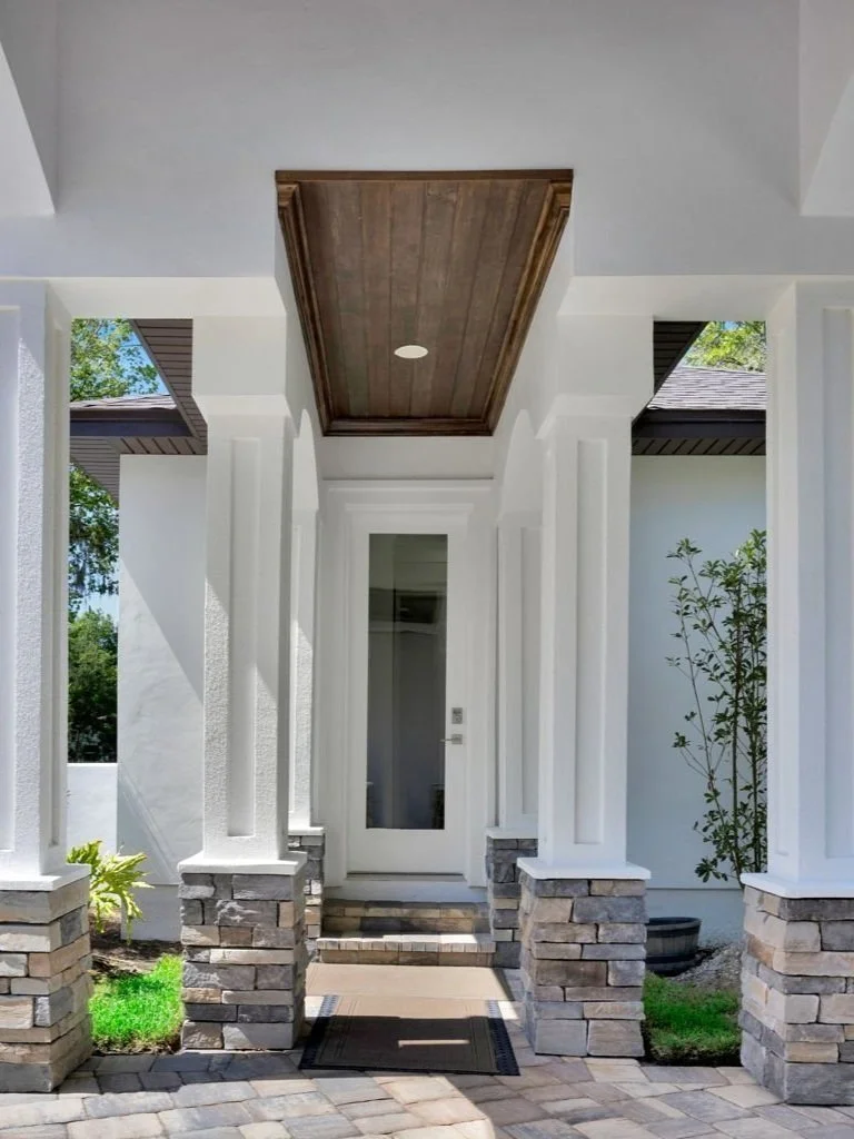 Front porch with white columns, stone bases, a wooden ceiling, and a glass door entrance. Green landscaping is visible on both sides.