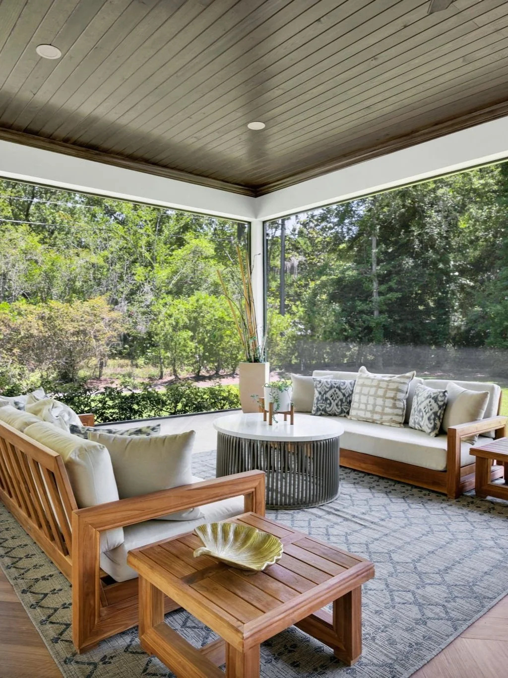 Sunroom with large windows overlooking lush green trees, beige sectional sofa with patterned pillows, wooden coffee table, and decorative plant in a white pot.