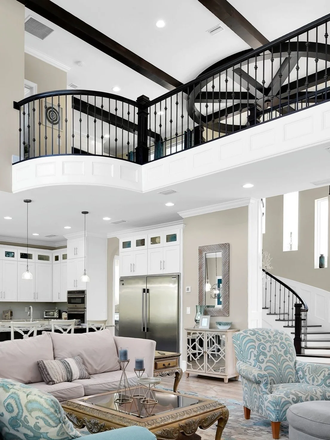 Interior view of a modern open-concept living area with a high ceiling, featuring a staircase with black railing, a kitchen with white cabinets, and a seating area with a beige sofa and patterned armchair.