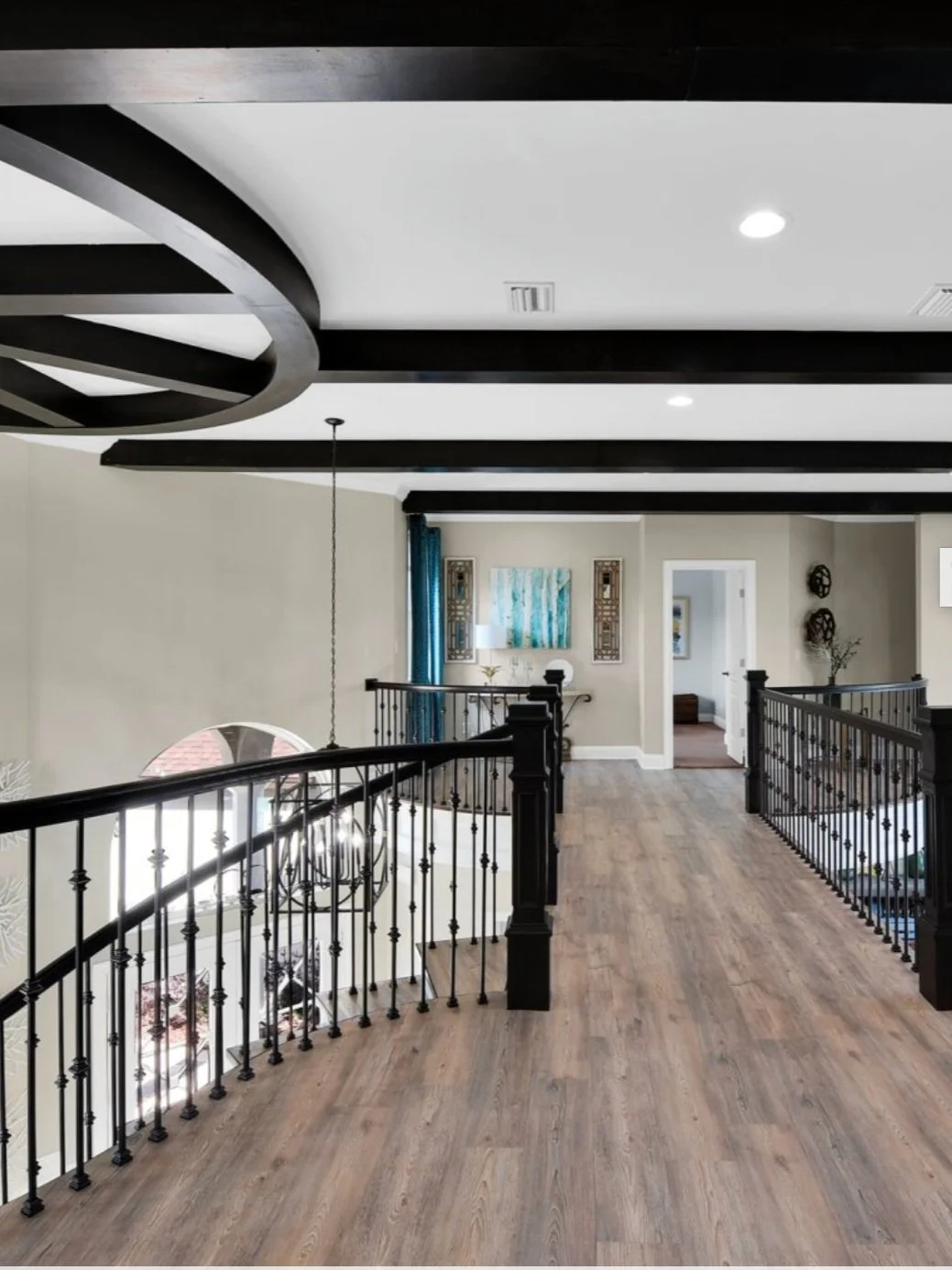 Interior view of a house upper floor with wood flooring, black railing, ceiling beams, and artwork on the walls.