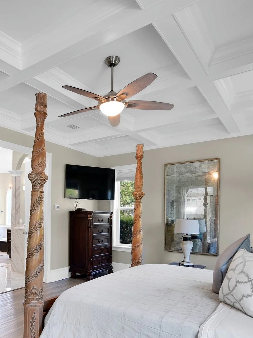 A bedroom featuring a white coffered ceiling, a ceiling fan with wooden blades, a flat screen TV mounted on the wall, a dark wooden dresser, a window with blinds, a large mirror, a bedside lamp, and a bed with a white quilt and pillow.