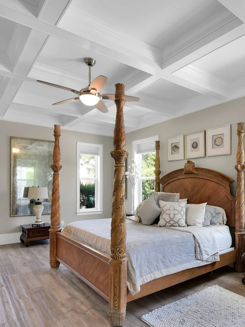 A bedroom featuring a wooden four-poster bed with intricate carved posts, a ceiling fan with lights, a bedside table with a lamp, and framed artwork on the wall.