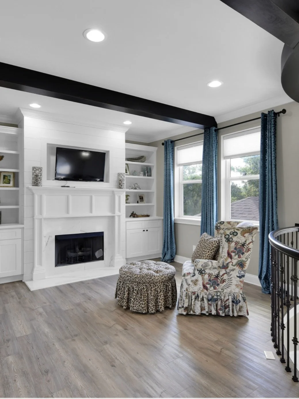 Living room with white built-in shelves, a white fireplace, a flat-screen TV, a floral armchair with a pillow, a leopard print ottoman, blue curtains, and windows overlooking trees.