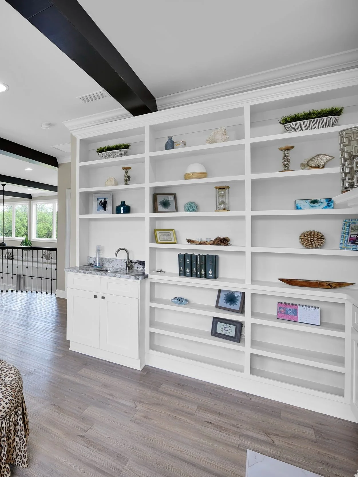 A white built-in bookshelf with various decorative items and a small marble sink with a faucet, inside a room with light wood flooring and a view of a living area with large windows.