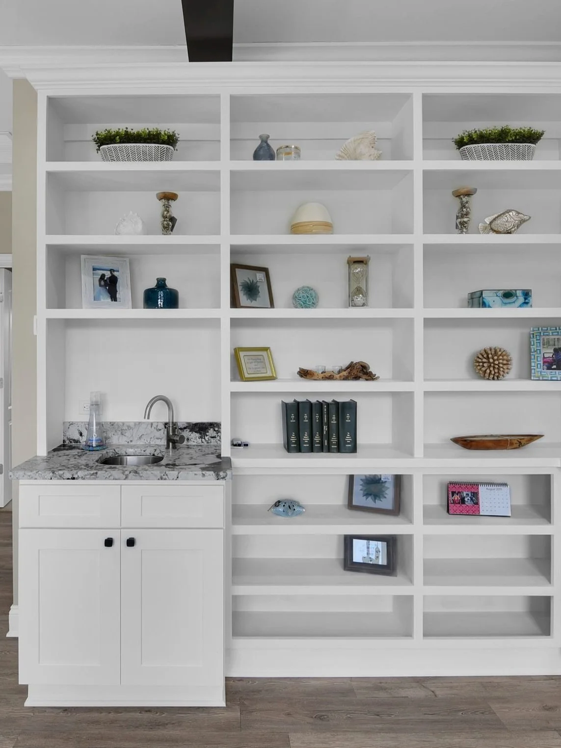 White built-in shelving unit with decorative objects, framed photos, books, and a small sink with a granite countertop at the lower left.