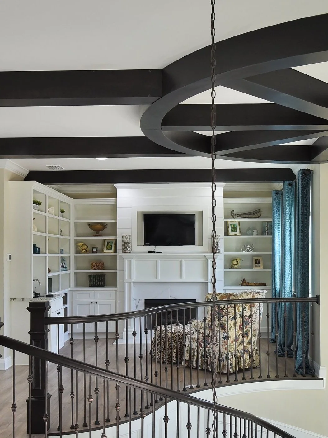 Living room with black ceiling beams, a black chandelier, and built-in white bookshelves around a white fireplace with a mounted TV. Two floral-patterned chairs and blue curtains are also visible.