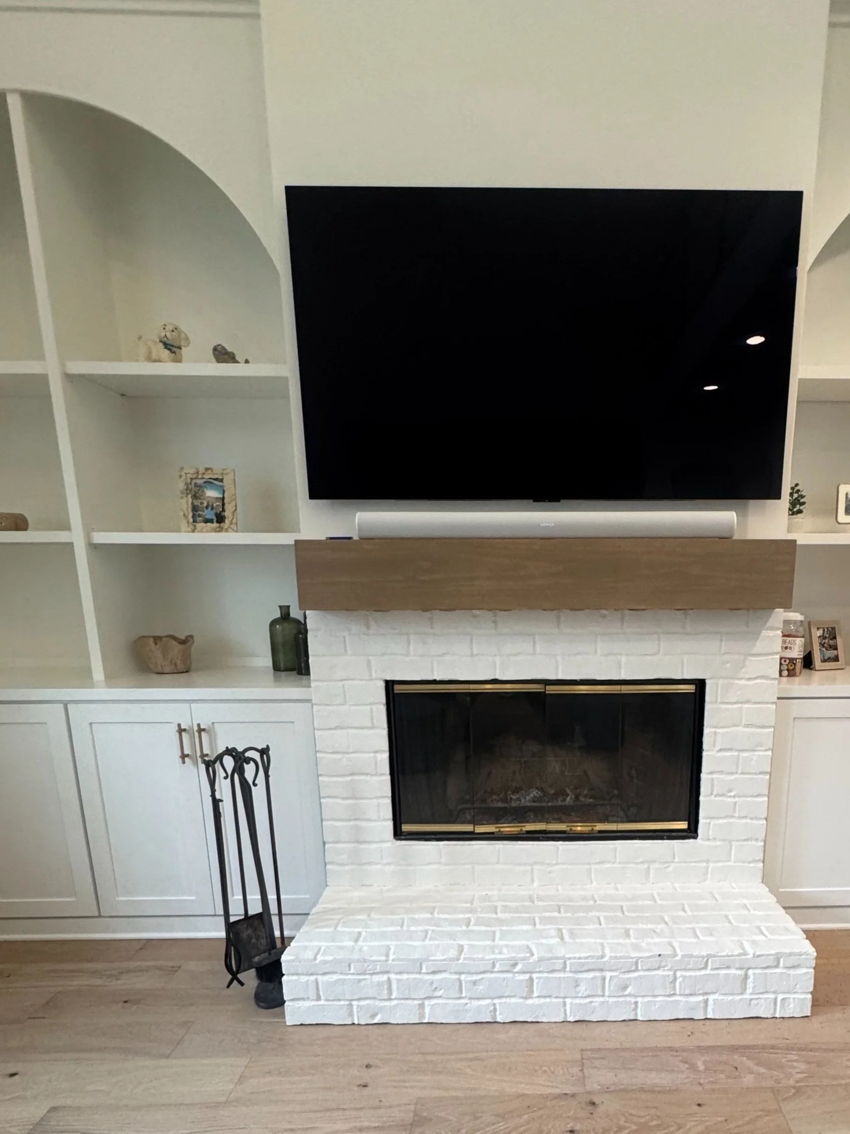 Living room fireplace with a mounted flat-screen TV above it. The fireplace is white brick with a wooden mantle. To the left of the fireplace are built-in shelves with decorative items and framed photos. A set of black fireplace tools is beside the f