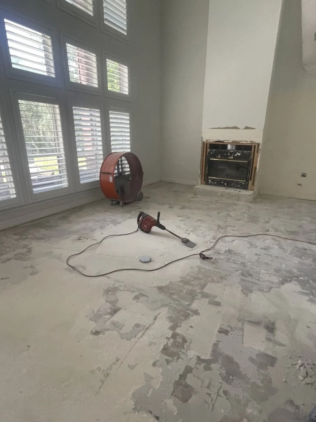Room under renovation with damaged flooring, large window with white shutters, a red air mover, a red-handled power tool on the floor, and an unfinished fireplace.