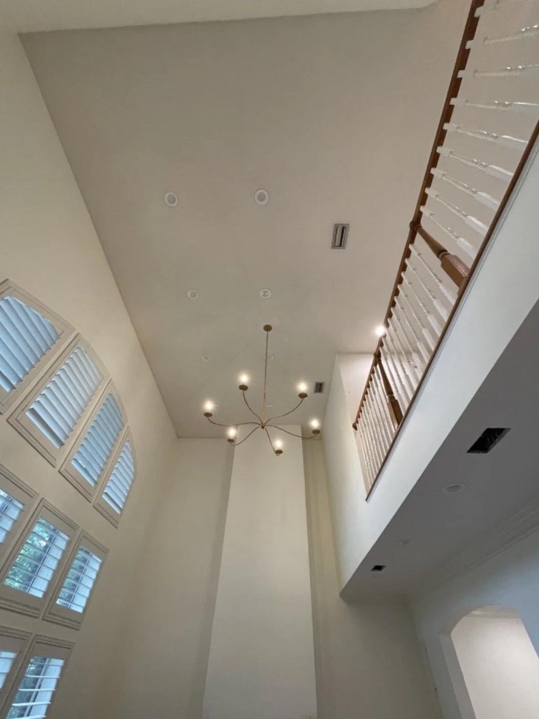 Interior view of a tall ceiling in a house with a modern chandelier, large arched windows with plantation shutters, a small balcony with wooden railing, and recessed lighting.