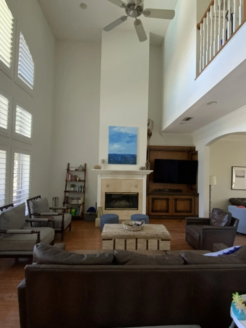 A spacious living room with high ceiling, large windows with blinds, a ceiling fan, and a fireplace with a piece of artwork above it. The room features a mix of old-fashioned and modern furniture, including armchairs, a sofa, a wooden coffee table, a