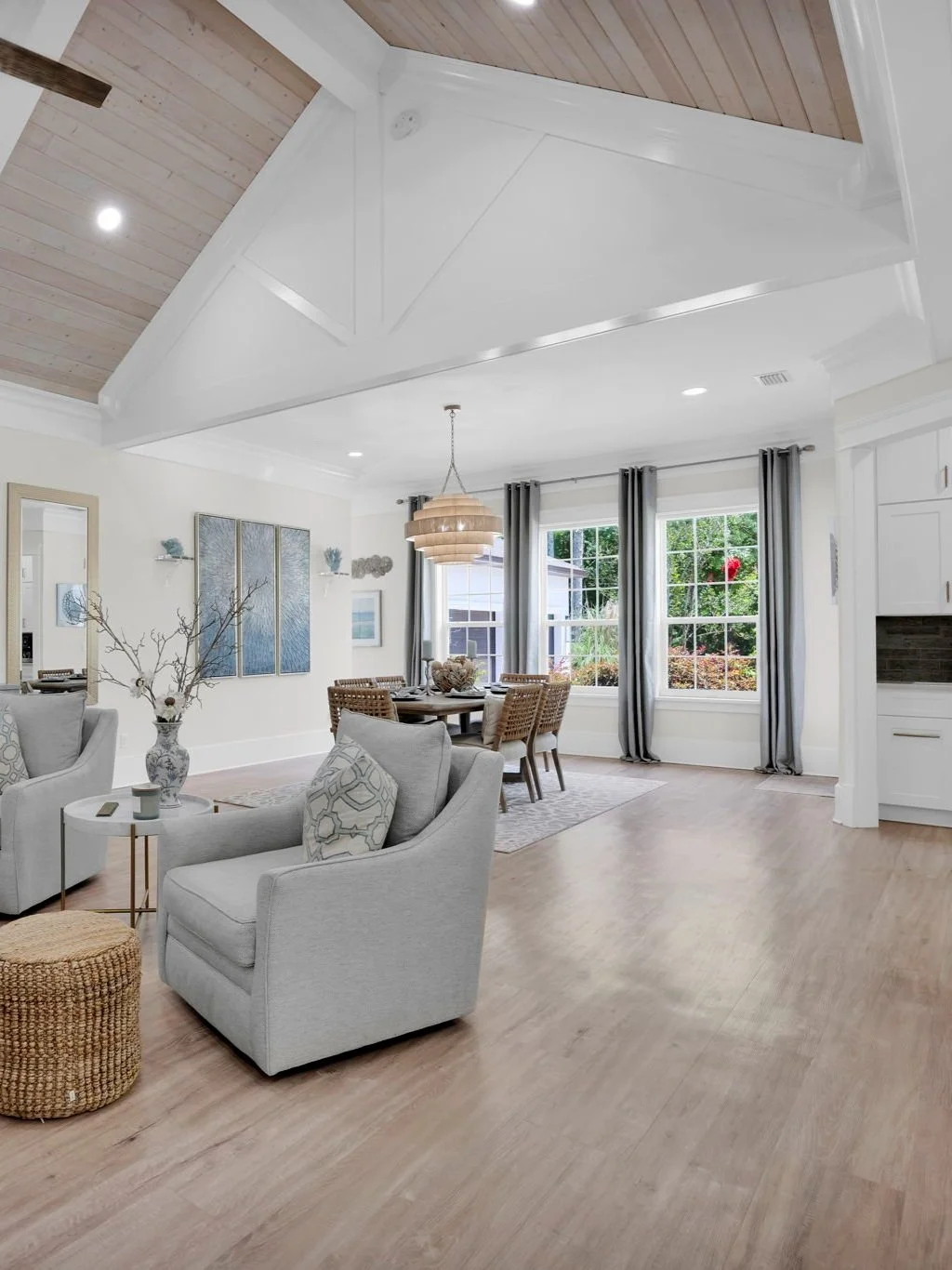 Bright, airy living and dining room with light wood floors, white walls, large windows with dark curtains, modern furniture including gray armchairs, a wicker dining table with chairs, artwork on the walls, a chandelier, and decorative items amid a m