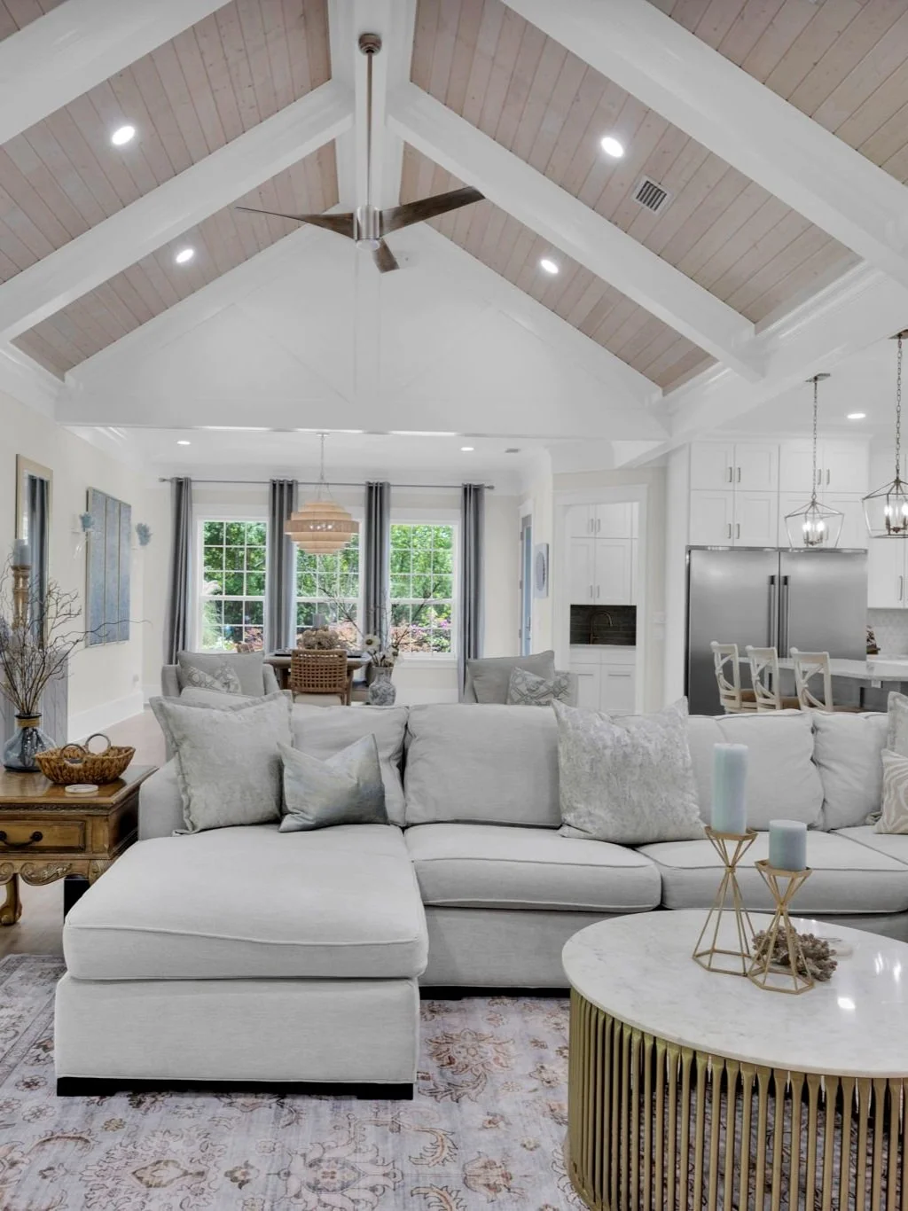 Bright living room with white sofa, decorative pillows, marble-topped coffee table, and natural light from large windows with gray curtains; open kitchen with stainless steel refrigerator and white cabinetry; vaulted ceiling with wood paneling and ce