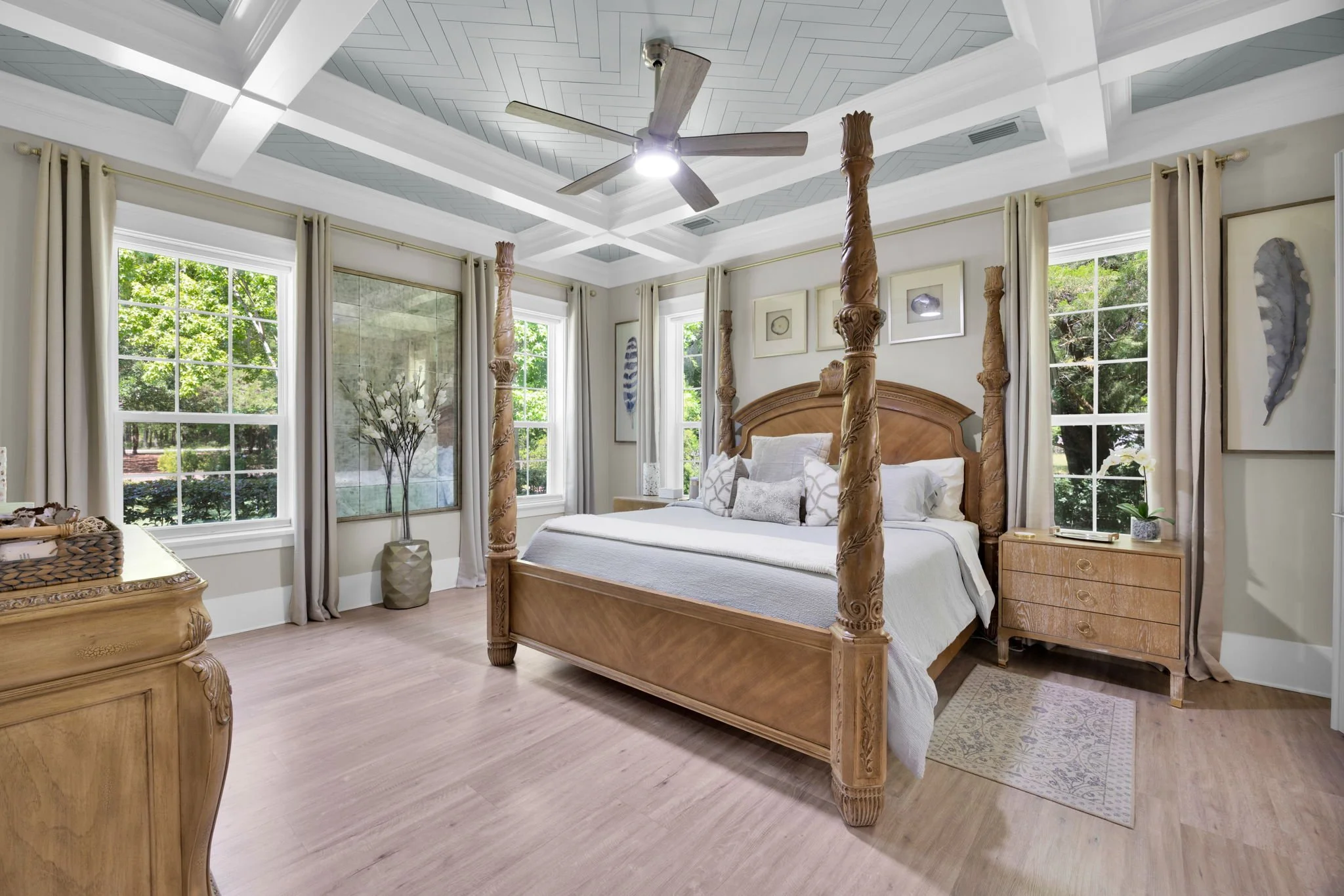 A bedroom with four large windows, beige curtains, a carved wooden four-poster bed with white bedding and pillows, a ceiling fan, a wooden dresser, a small wooden nightstand, a tall vase with flowers, and framed artwork on the walls.