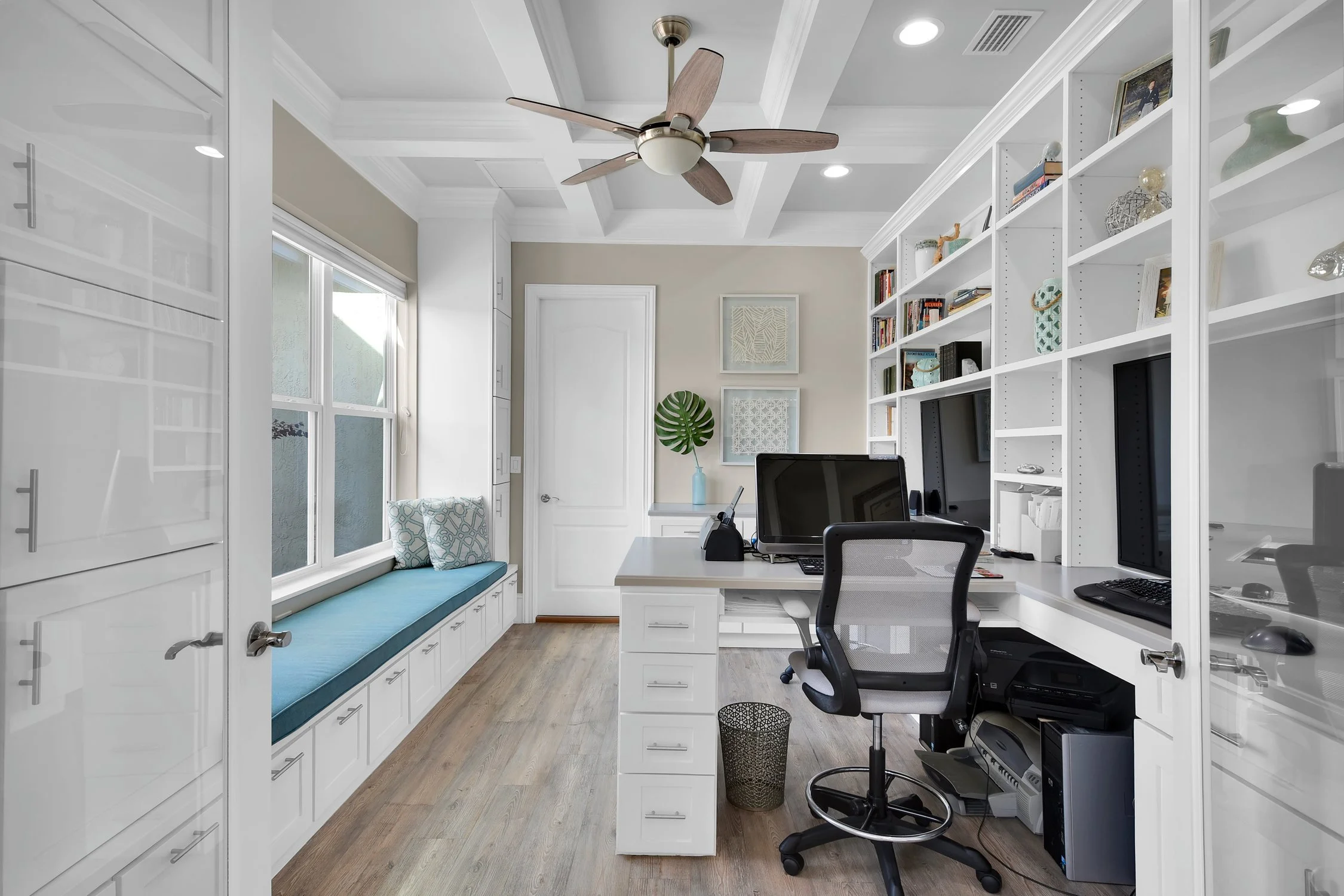 Home office with built-in white desk and shelves, black office chair, computer monitor, window with window seat and cushions, ceiling fan, and decorative wall art.
