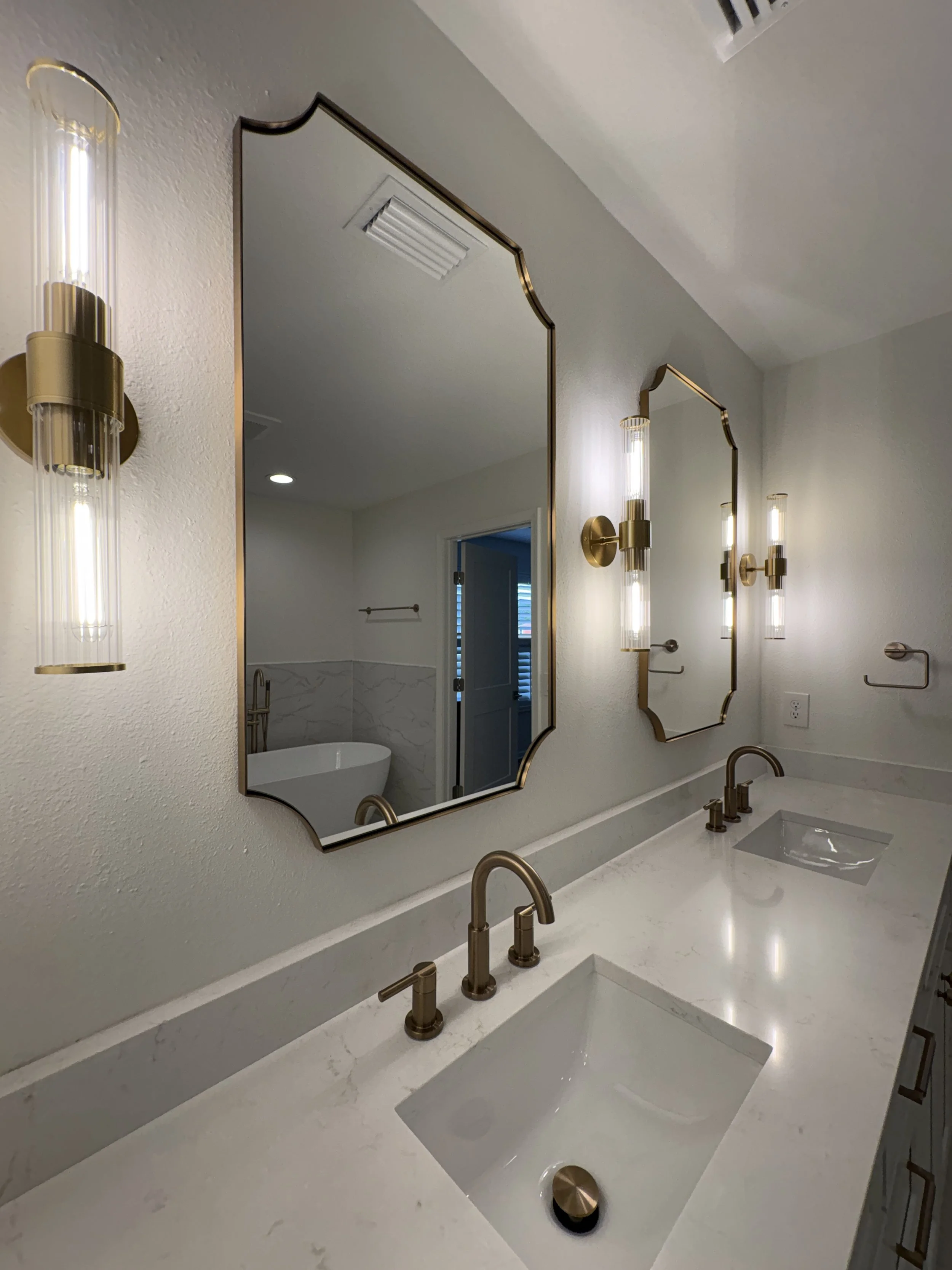 A custom double vanity with extensive storage, a sleek white countertop, decorative mirrors, and brushed gold fixtures brings elegance and function to this luxury Jacksonville condo bathroom remodel by Elite Construction of JAX.