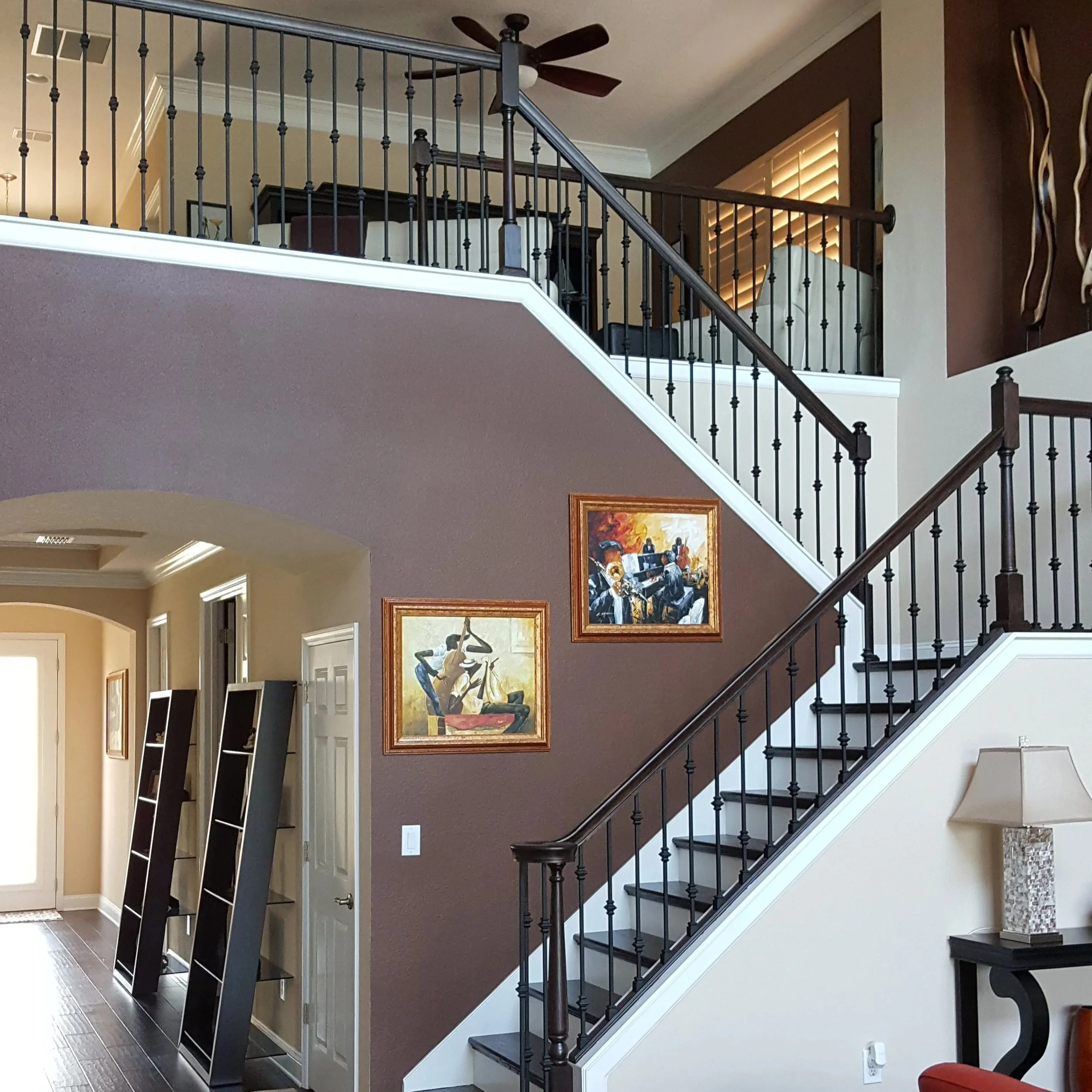 View of a modern staircase with black metal railing and white steps, artwork on brown wall, and a ceiling fan in a well-lit living area.