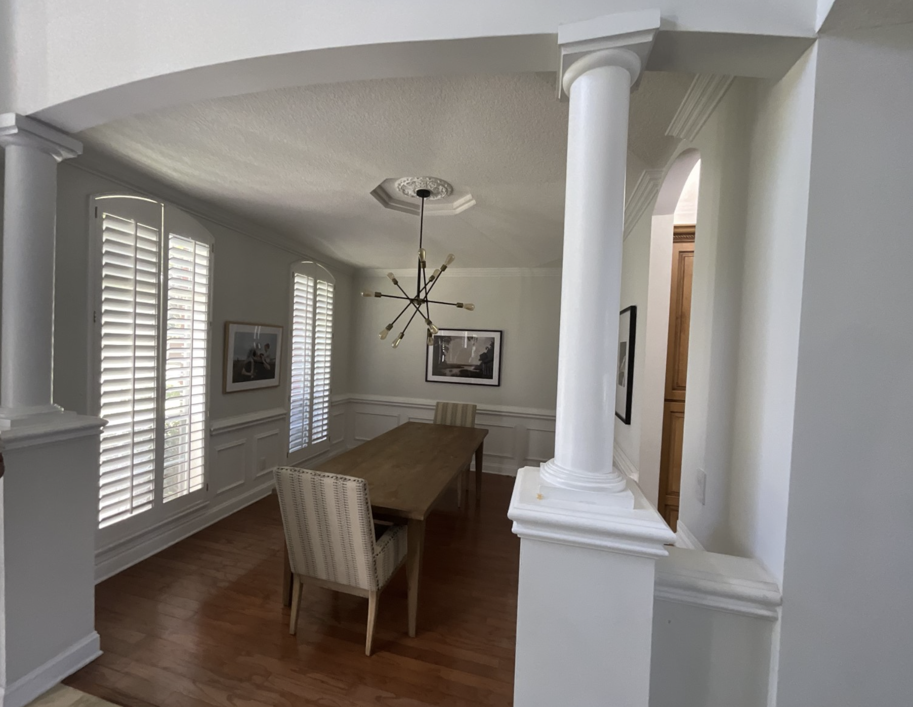 Dining room with a wooden table and a beige upholstered chair, white walls with wainscoting, large arched windows with shutters, and a modern chandelier hanging from the ceiling.