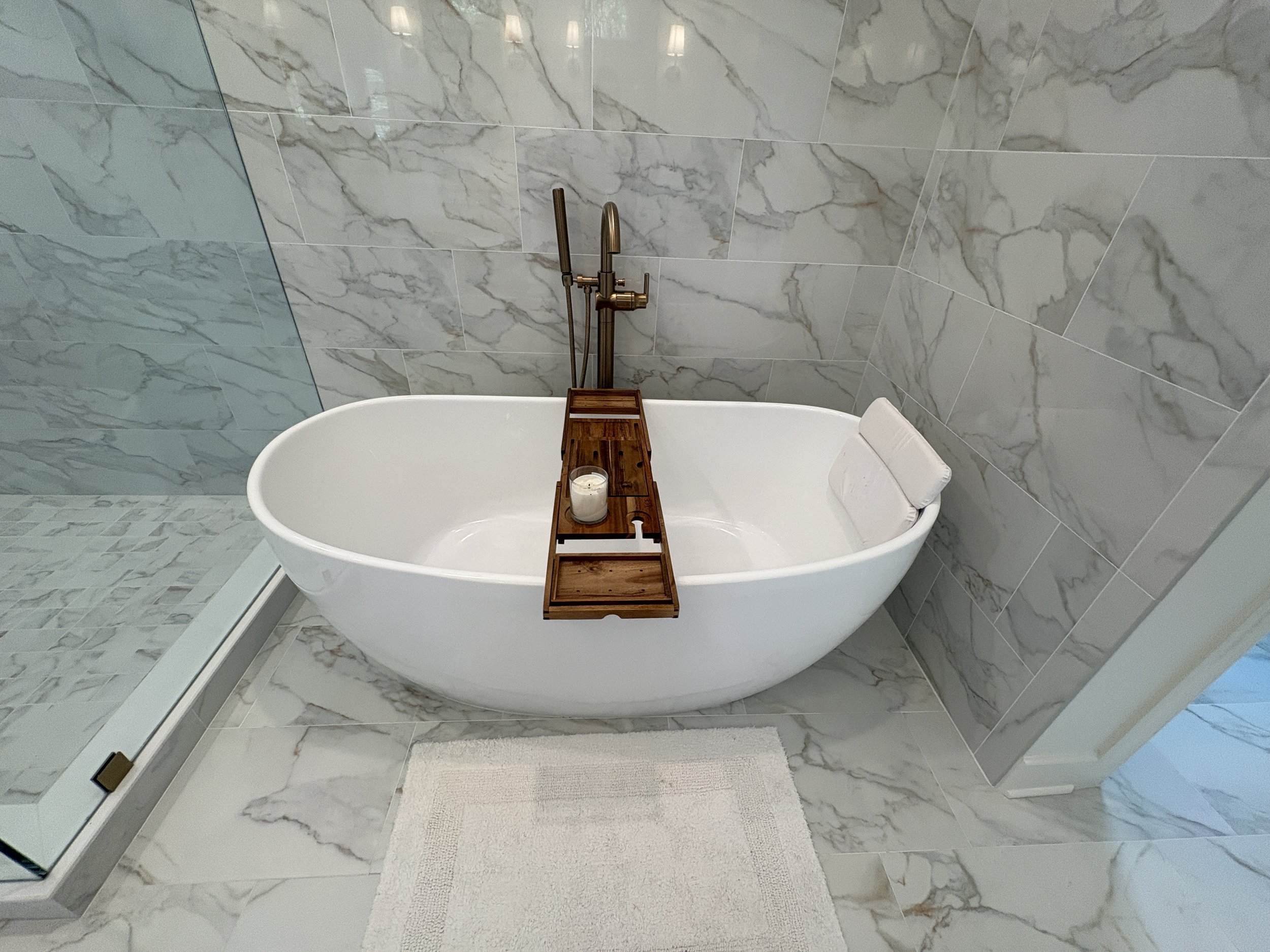 Finished primary bathroom featuring a freestanding soaking tub with a floor-mounted bronze filler, surrounded by large-format marble-look tile that creates a clean, luxurious aesthetic. The elegant tub alcove serves as a relaxing focal point, while p