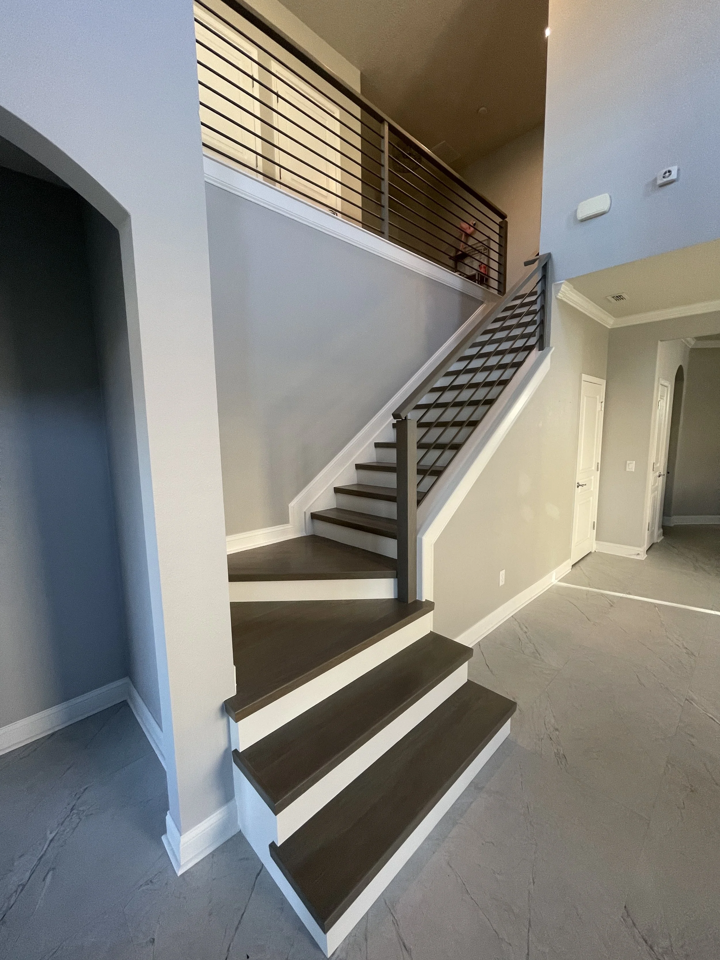 Finished modern stair renovation featuring wood treads with contrasting white risers and a custom horizontal metal railing system extending to the upper landing. Clean wall finishes, precise stair geometry, and coordinated flooring transitions emphas