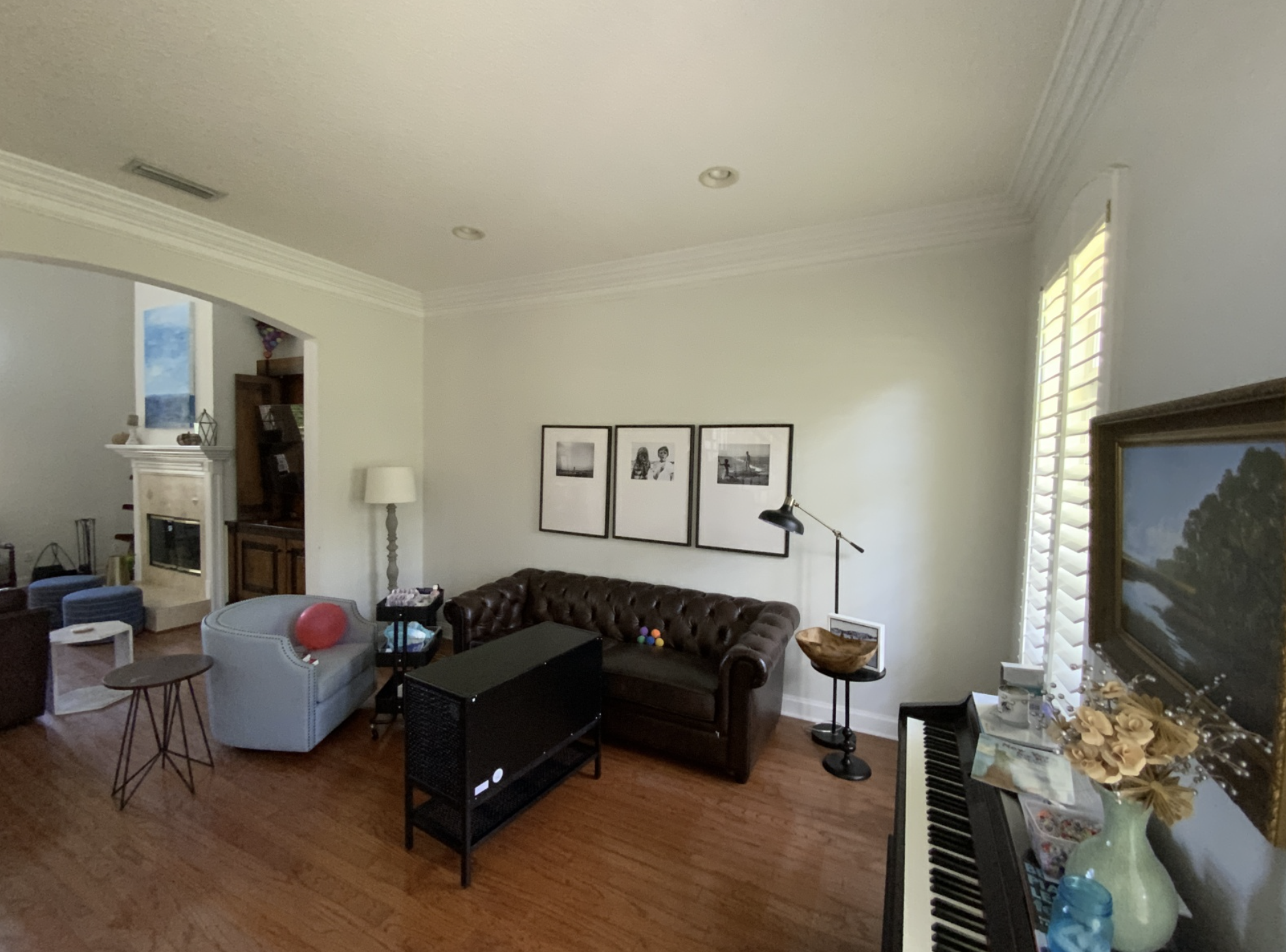 Living room with a brown leather couch, a white armchair, a small side table with a balloon, framed photos on the wall, a floor lamp, a piano with flowers and photos, and a large window with white shutters.