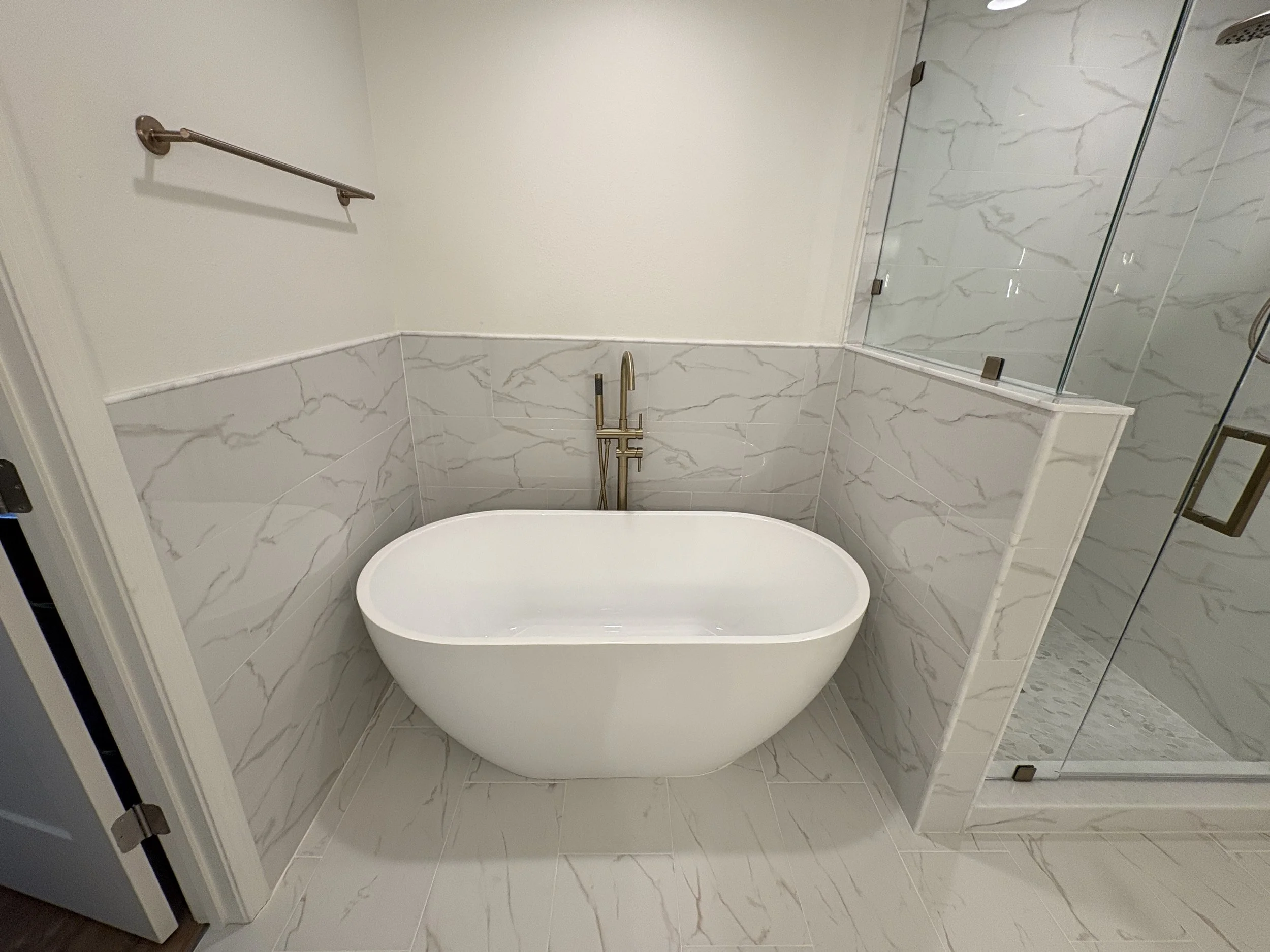A sculptural freestanding tub with brushed gold fixtures and marble-look tile creates a relaxing focal point in this luxury primary bathroom. This high-end Jacksonville condo remodel was completed by Elite Construction of JAX.