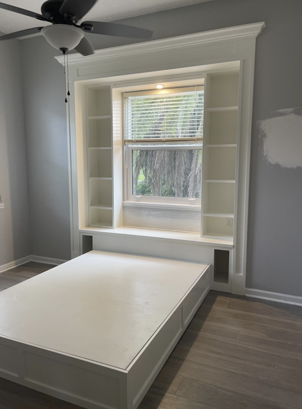 White built-in bed frame with side shelves under a window, in a room with gray walls, wooden flooring, and a ceiling fan.