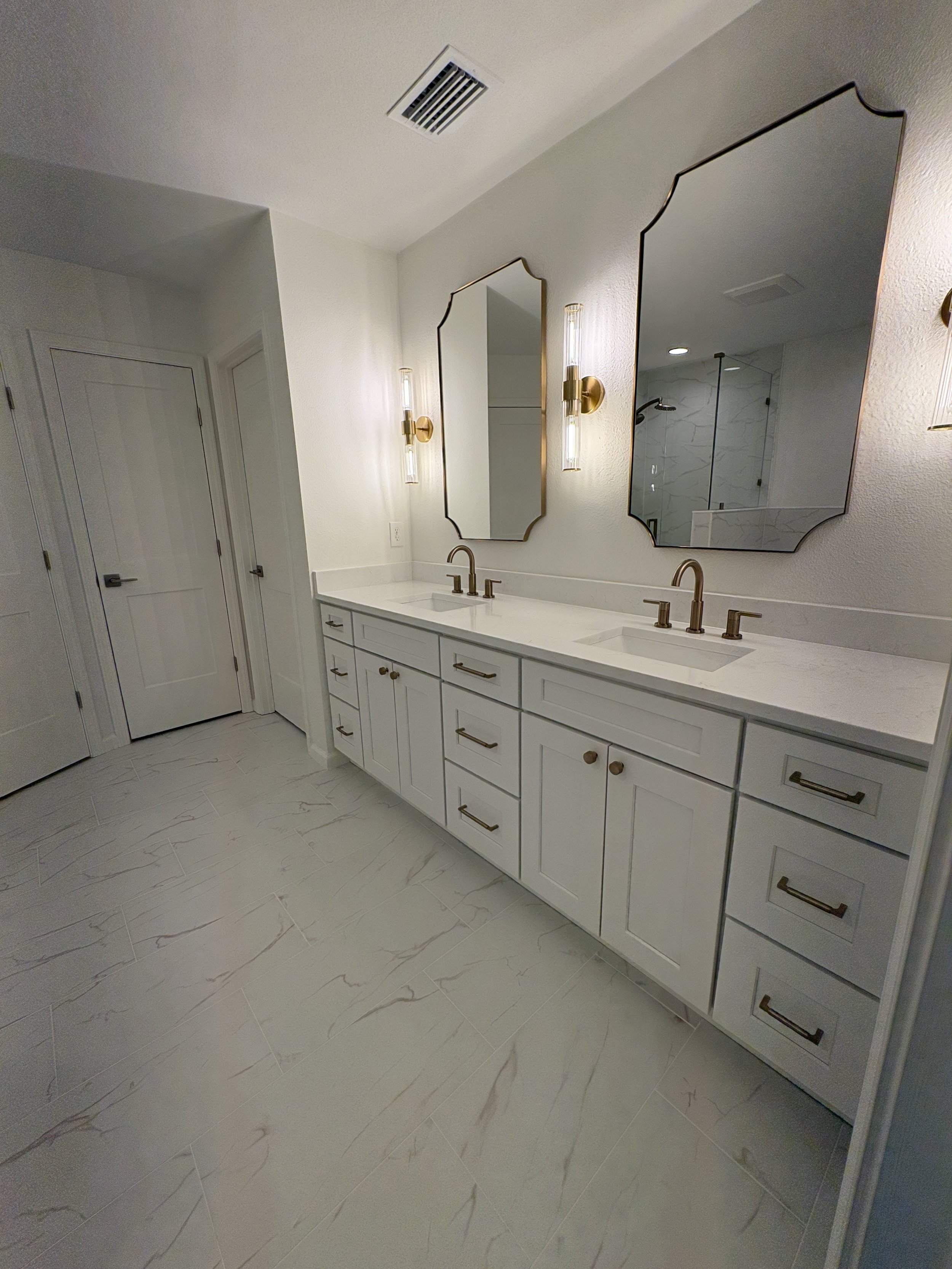 This luxury primary bathroom features a custom double vanity, full-height cabinetry, marble-look countertops and flooring, decorative mirrors, and brushed gold fixtures. Elite Construction of JAX completed this high-end condo remodel in Jacksonville,