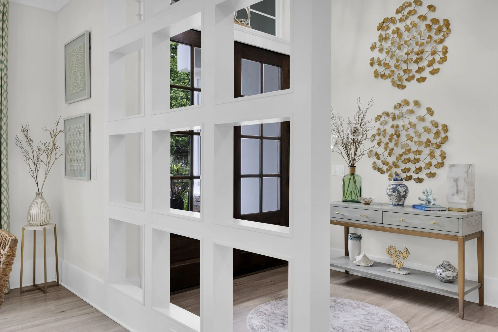 Interior of a modern home with a decorative white partition featuring square cutouts, wood and white framed windows, and a wall with gold ginkgo leaf wall art. There is a side table with a vase and dried branches, and a console table with decorative