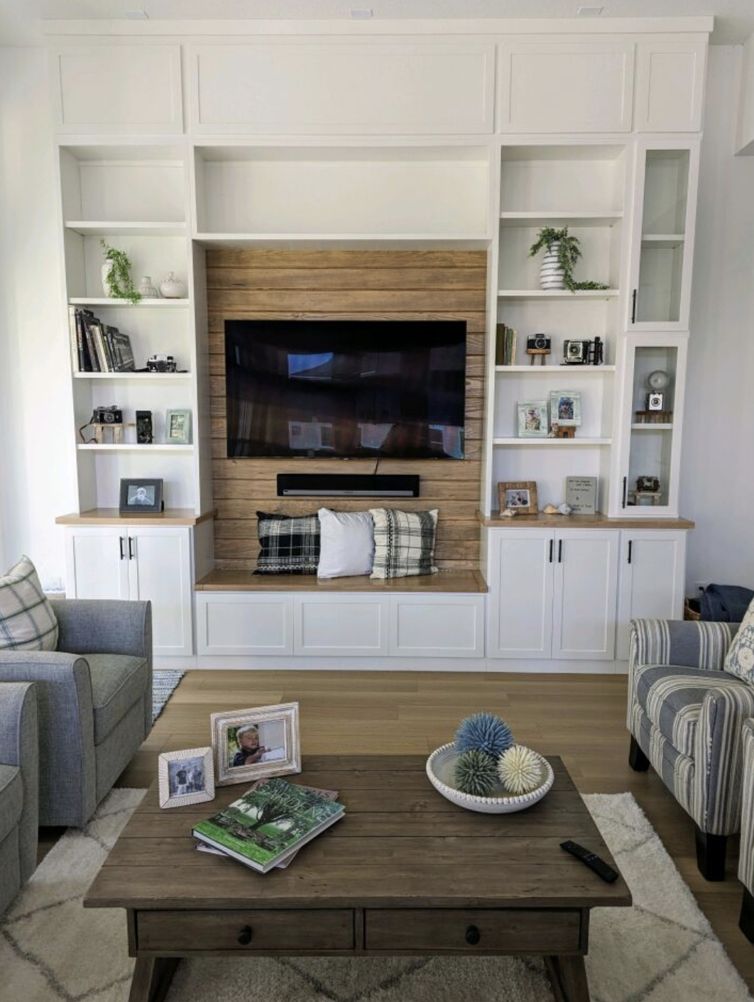Living room with a wall-mounted TV on a wooden panel, white built-in shelves with books, photos, and decorative items, a gray armchair with plaid pillow, a striped armchair, wooden coffee table with framed photos, a bowl of decorative balls, and remo