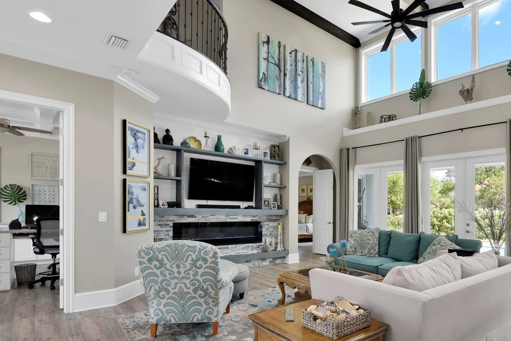 Bright living room with high ceilings, large windows, and a ceiling fan. Features a sectional sofa, an armchair, a coffee table, and a modern fireplace with a TV above. Decor includes artwork, plants, and shelves with decorative items.