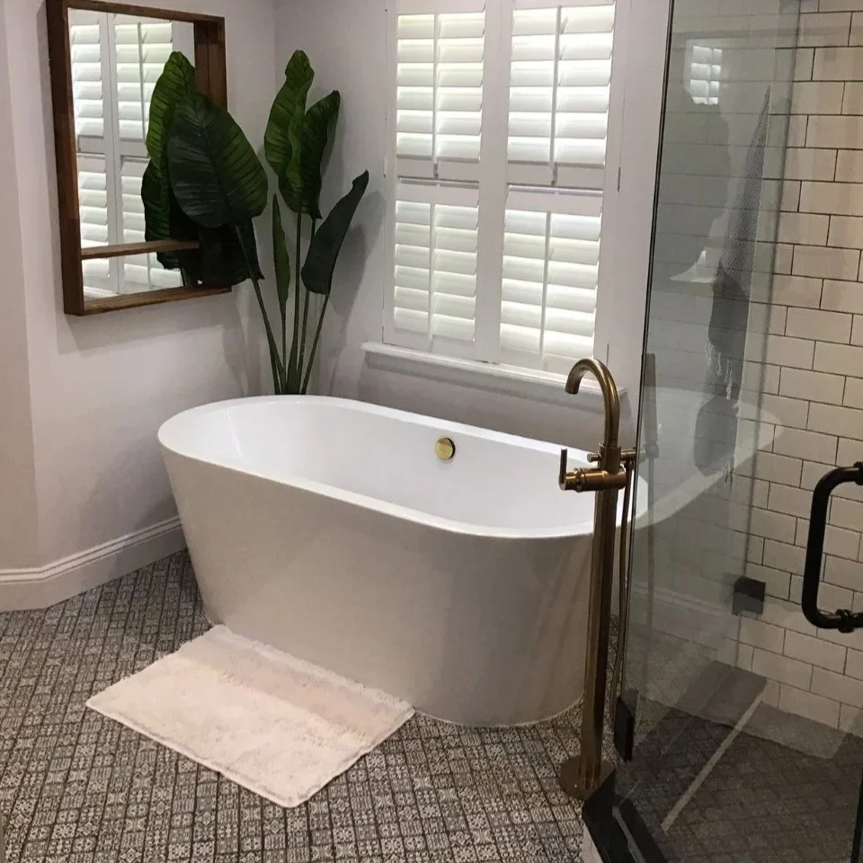 A modern bathroom with a white freestanding bathtub, a large leafy green plant, white plantation shutters on the window, and a glass shower door with a gold handle.