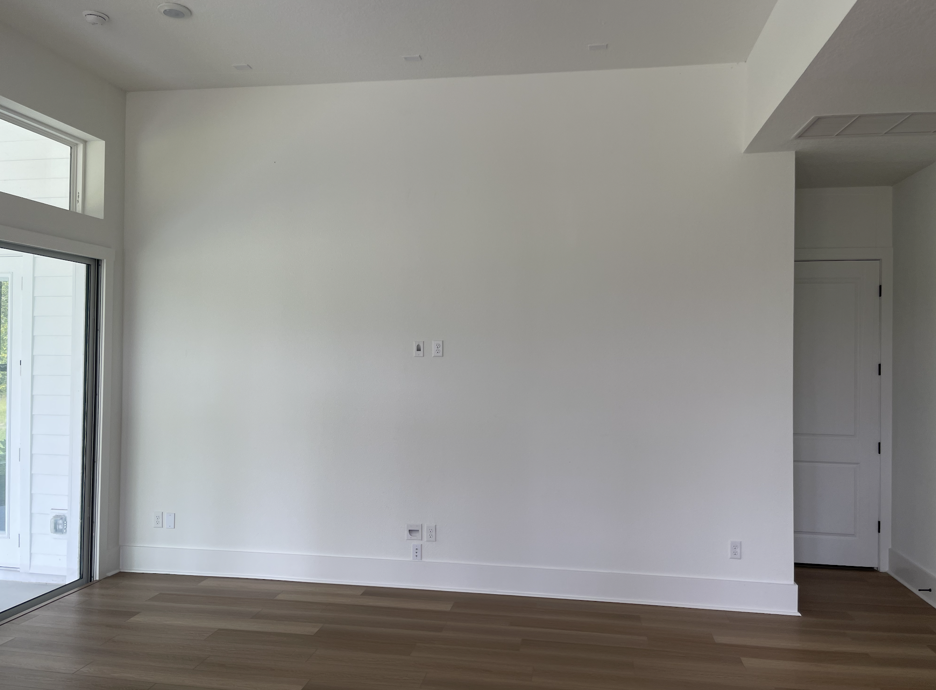 Empty white room with hardwood floors, a large sliding glass door, and electrical outlets on the wall.