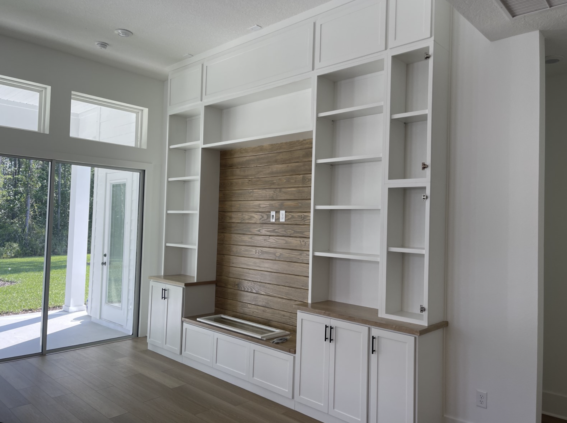 Empty built-in white shelves and cabinets with a wood panel accent wall in a bright room, next to sliding glass doors leading outside.