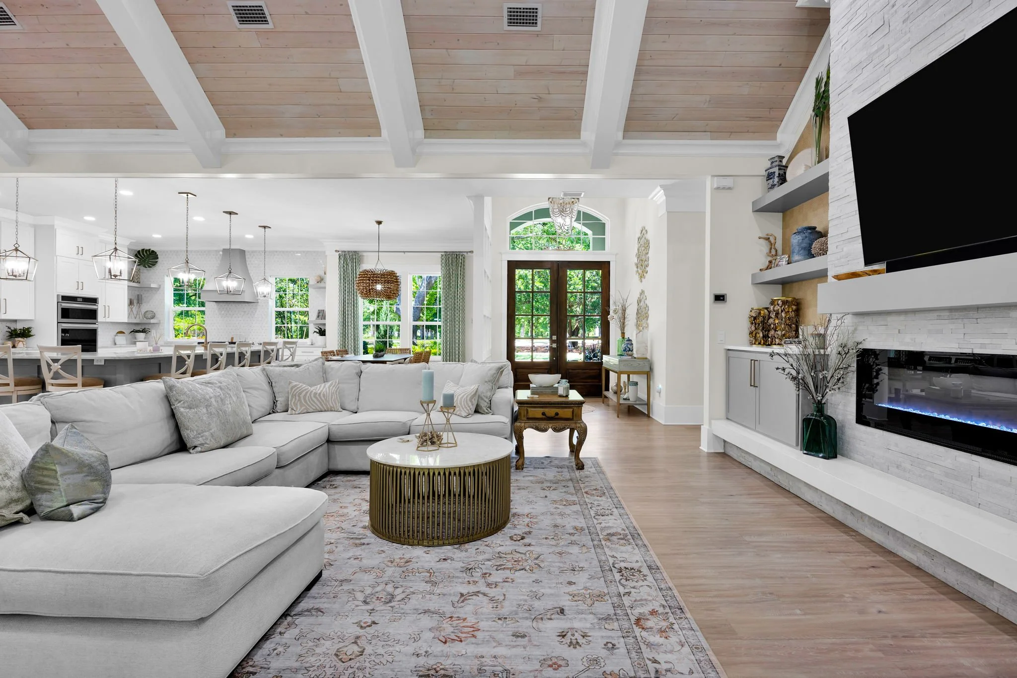 Spacious living room with white sectional sofa, round marble coffee table, and decorative pillows. Open kitchen with white cabinets and pendant lighting in the background. Large glass front door, hardwood floors, and a built-in fireplace with shelves