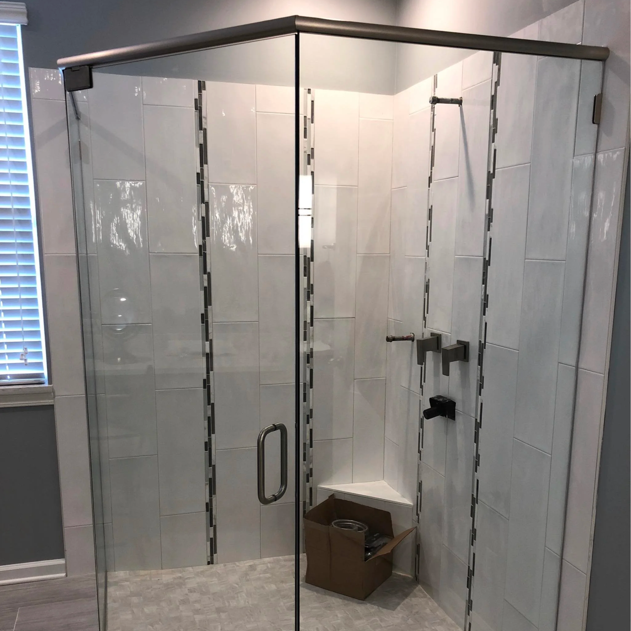 A partially assembled shower in a bathroom with white tiled walls and gray flooring. The glass door is installed, but the shower fixtures are not yet mounted, and there is an open cardboard box at the corner.