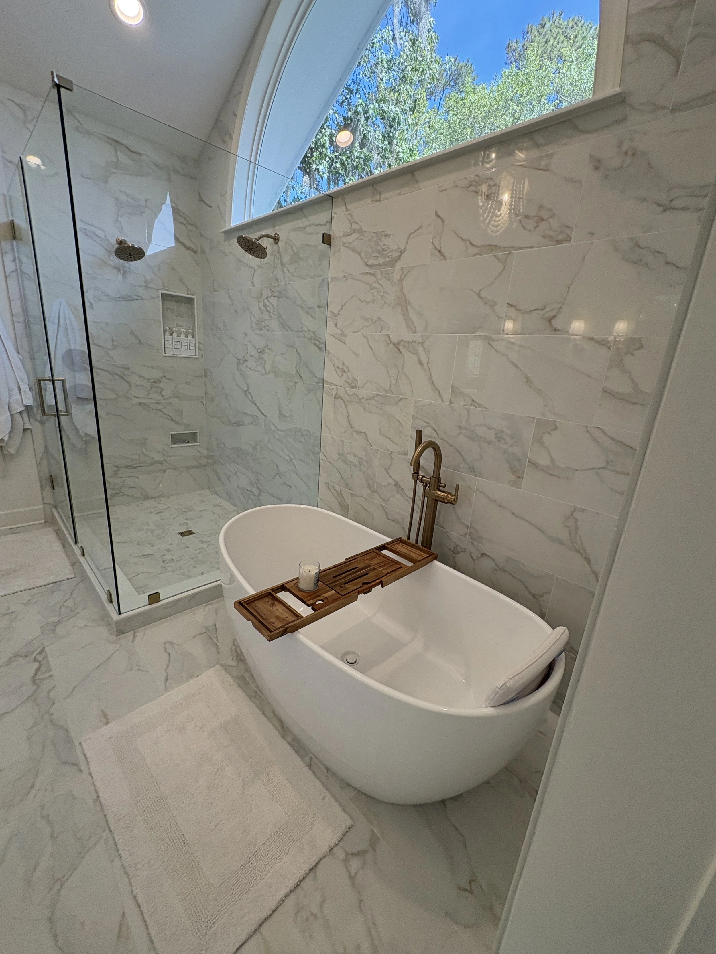 Finished primary bathroom featuring a freestanding soaking tub, frameless glass walk-in shower, and floor-to-ceiling marble-look tile that creates a bright, open, and luxurious atmosphere. An oversized arched window fills the space with natural light