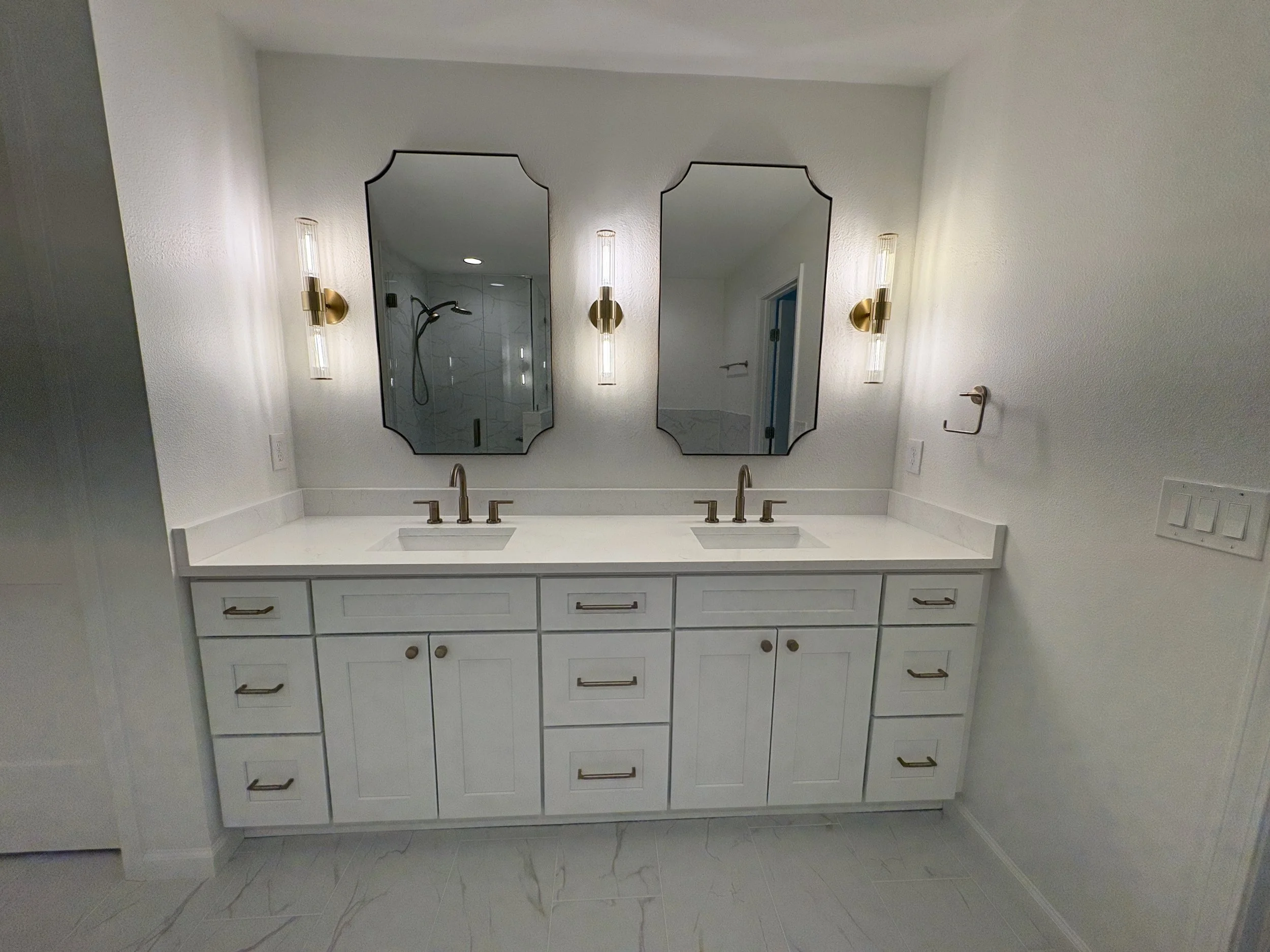 A custom double vanity with extensive storage, a sleek white countertop, decorative mirrors, and brushed gold fixtures brings elegance and function to this luxury Jacksonville condo bathroom remodel by Elite Construction of JAX.