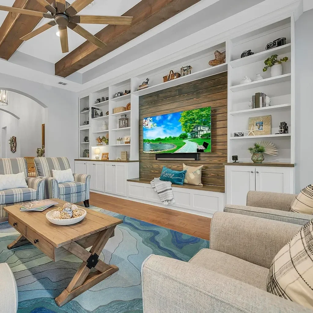 A cozy living room with a wood-paneled accent wall and a large flat-screen TV. The room includes white built-in shelves, a beige armchair, striped armchairs, a wooden coffee table, and a colorful area rug. The ceiling features wooden beams and a ceiling fan.