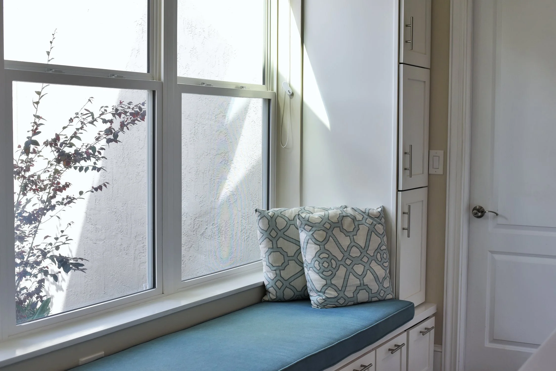 A window seat with a blue cushioned bench and two patterned pillows, next to a white wall with cabinets and a door.