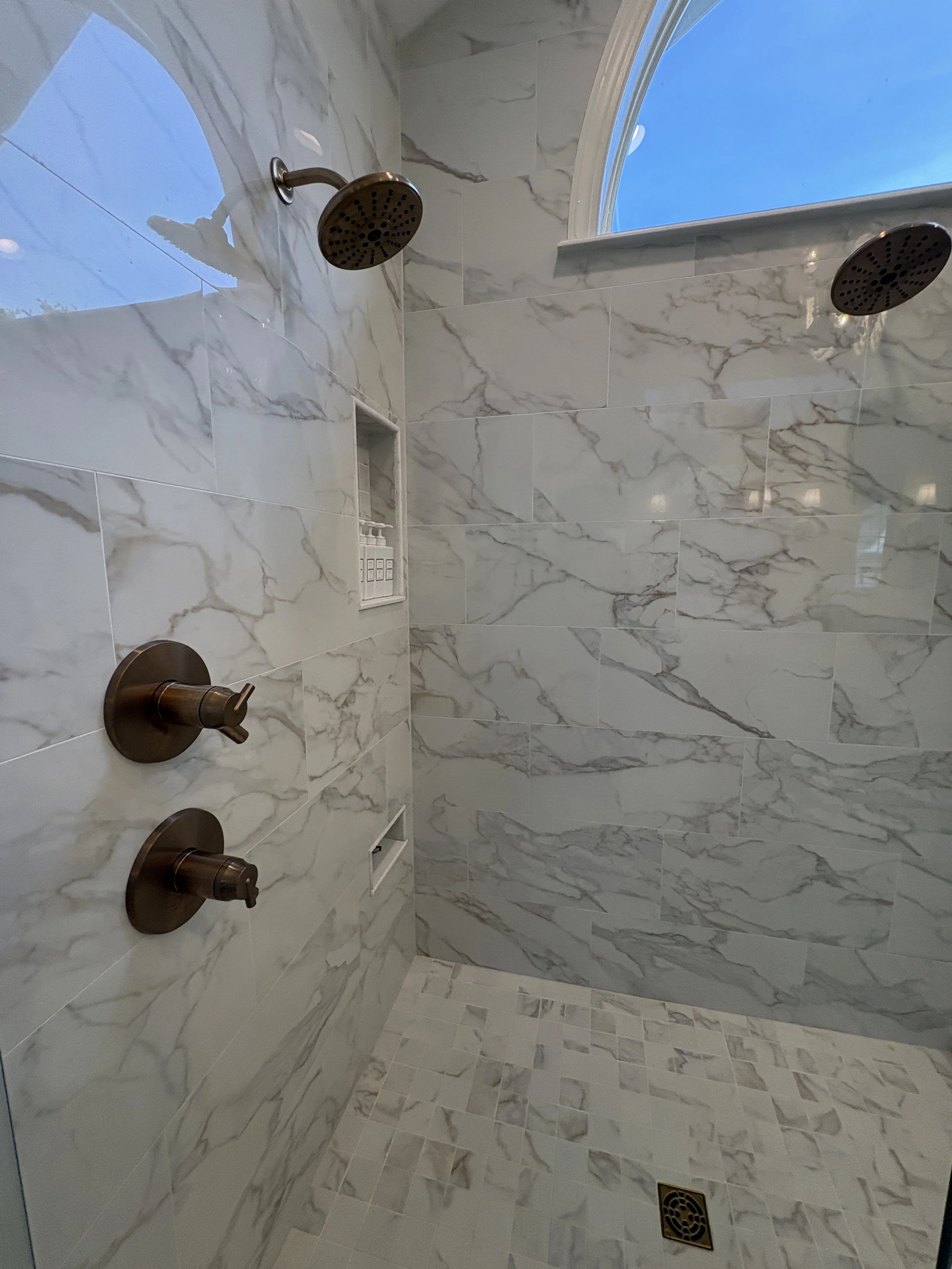 Finished walk-in shower featuring a frameless glass enclosure, oversized marble-look wall tile, and a custom tiled shower pan designed for a seamless, open-concept appearance. Natural light from the dramatic arched window enhances the bright, spa-ins