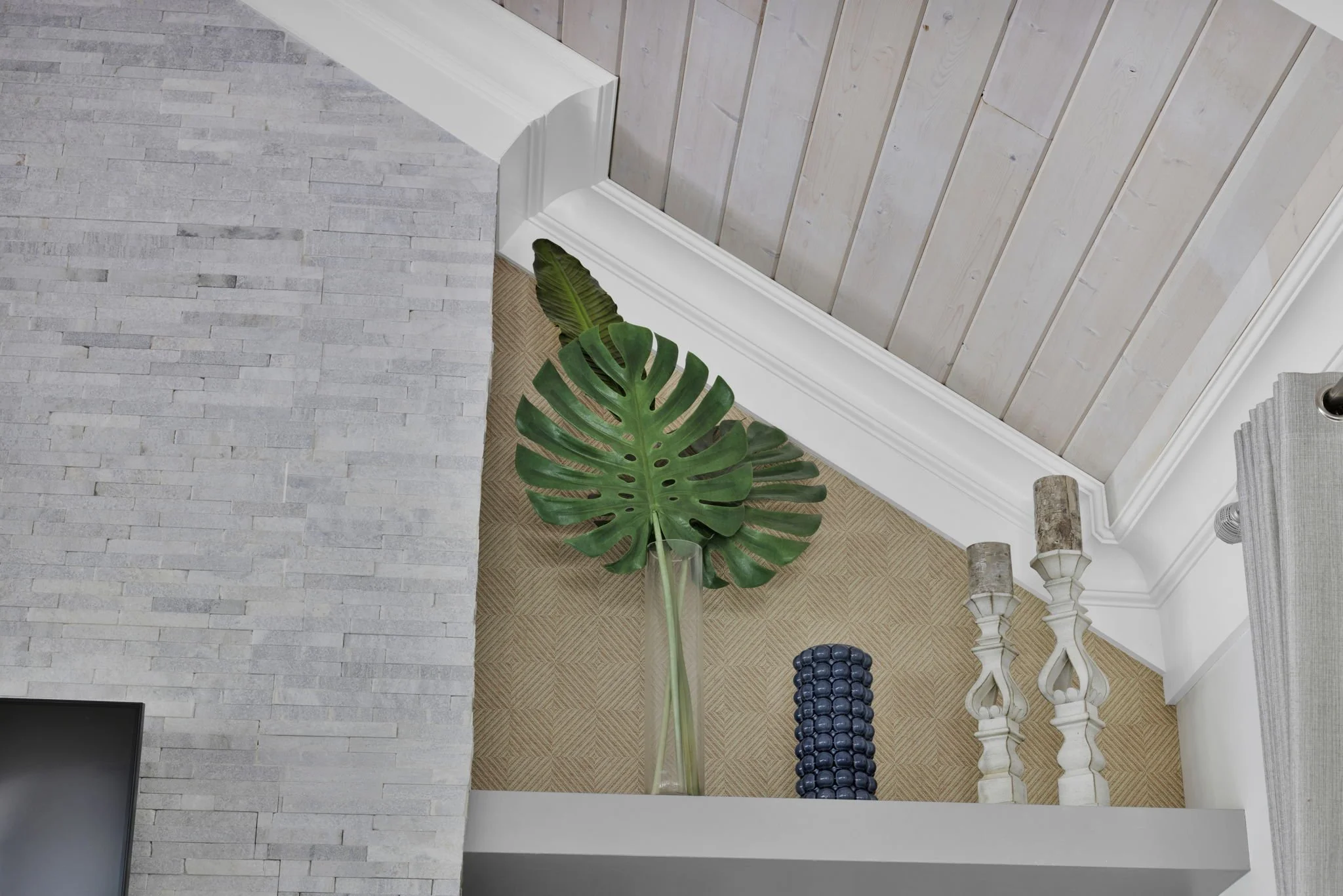 Interior upper corner of a living room with gray brick fireplace, white wainscoting, beige carpet, and decorative items including a large green monstera leaf in a glass vase, a cylindrical blue sculpture, and two candle holders.