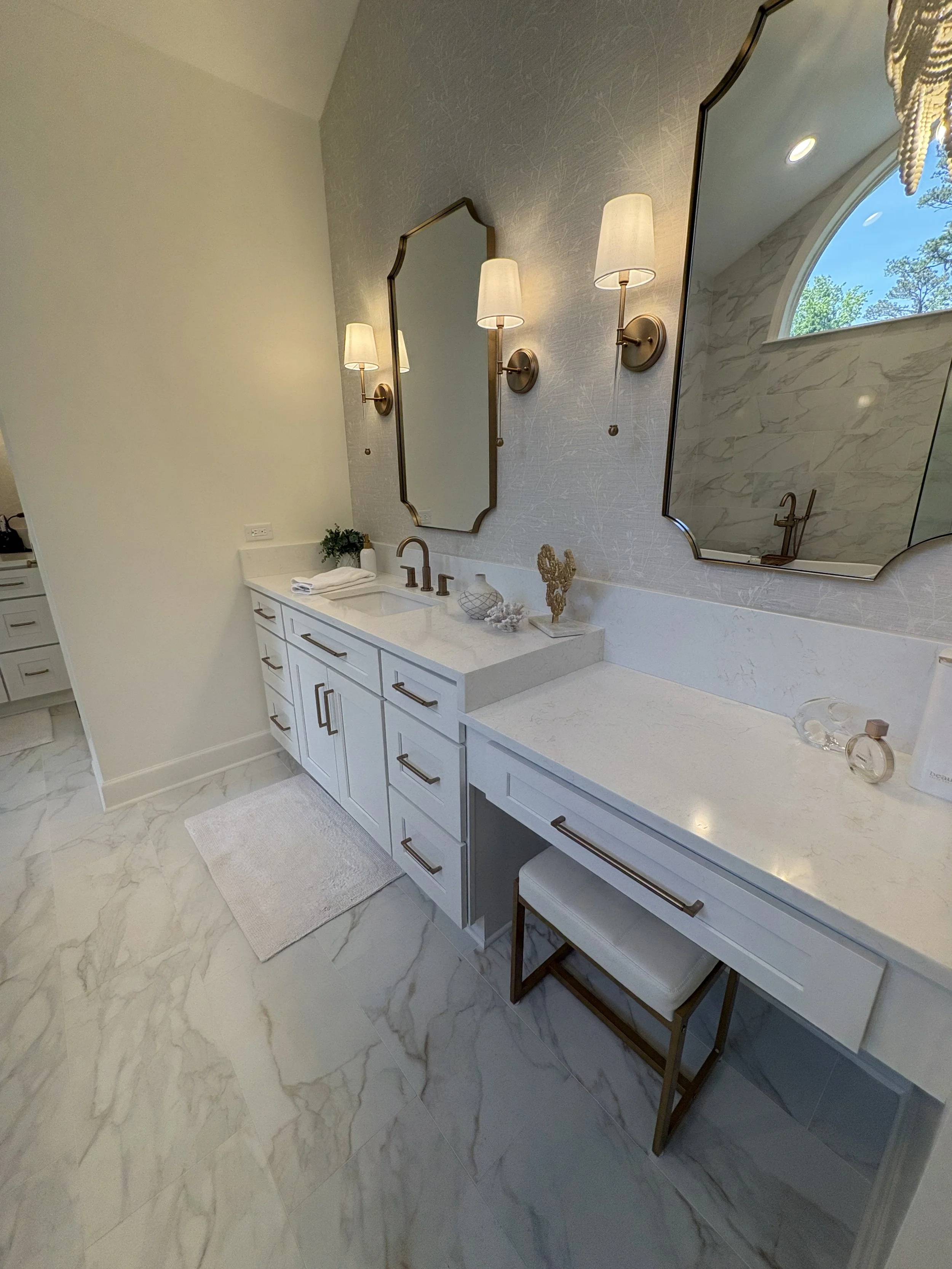 Finished primary bathroom vanity area featuring custom white shaker cabinetry, quartz countertops, dual undermount sinks, and a dedicated makeup station designed for everyday convenience. Decorative mirrors, elegant wall sconces, and refined hardware