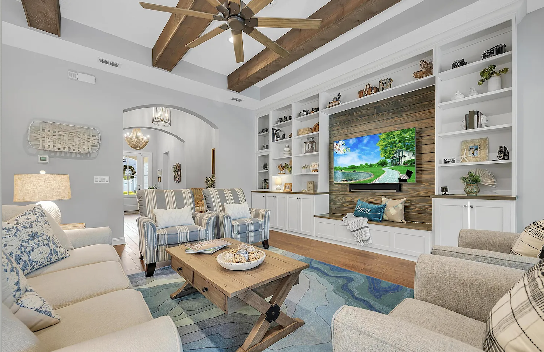 Living room with a TV mounted on a wooden-paneled wall, surrounded by white built-in shelves filled with decorative items and cameras. There are beige and striped armchairs, a sofa with patterned pillows, a wooden coffee table, a colorful area rug, a
