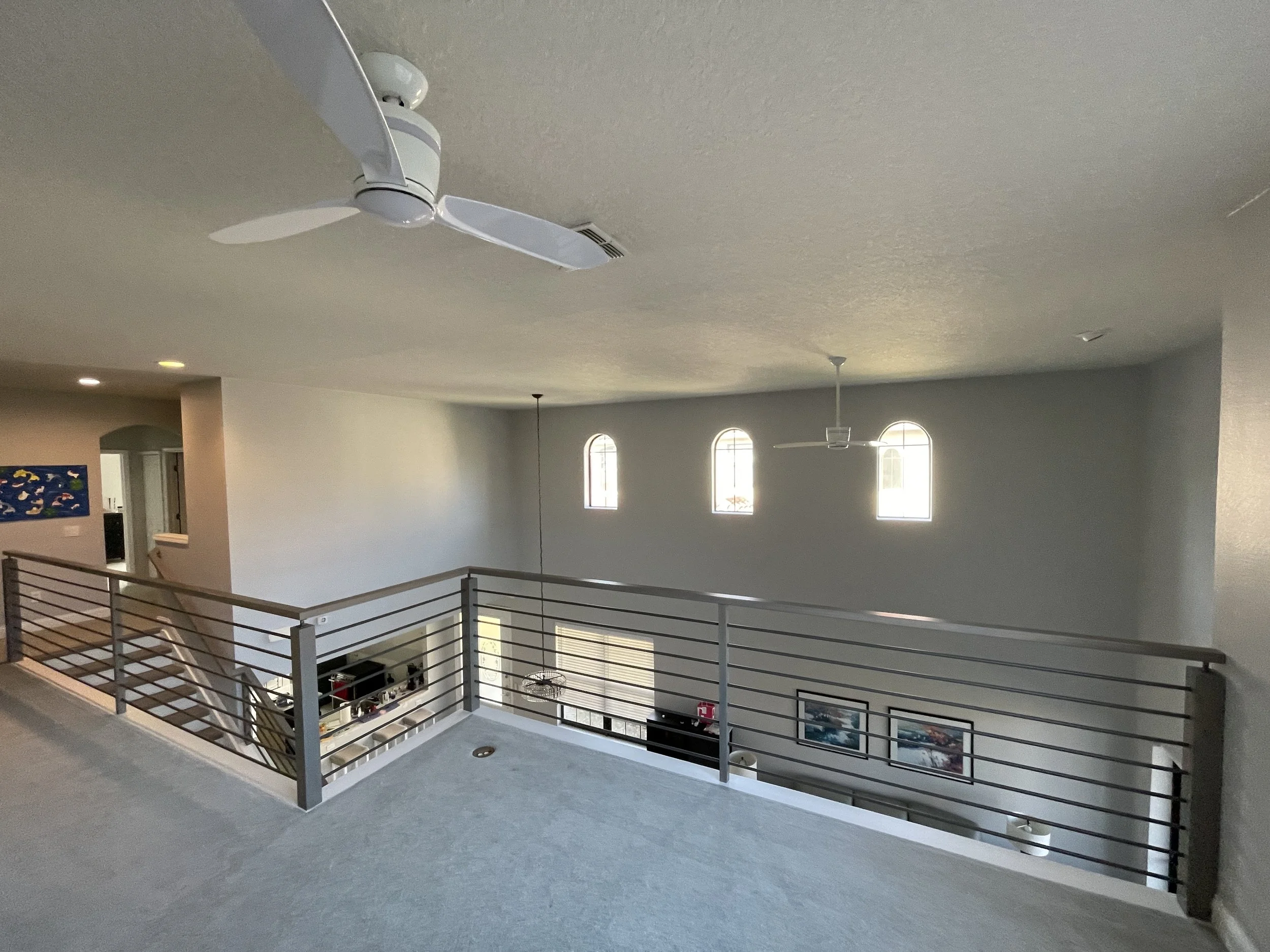 Finished upper-level loft renovation featuring a custom horizontal metal railing system overlooking the living space below. Clean wall finishes, integrated lighting, and a modern ceiling fan installation create an open, functional secondary living ar