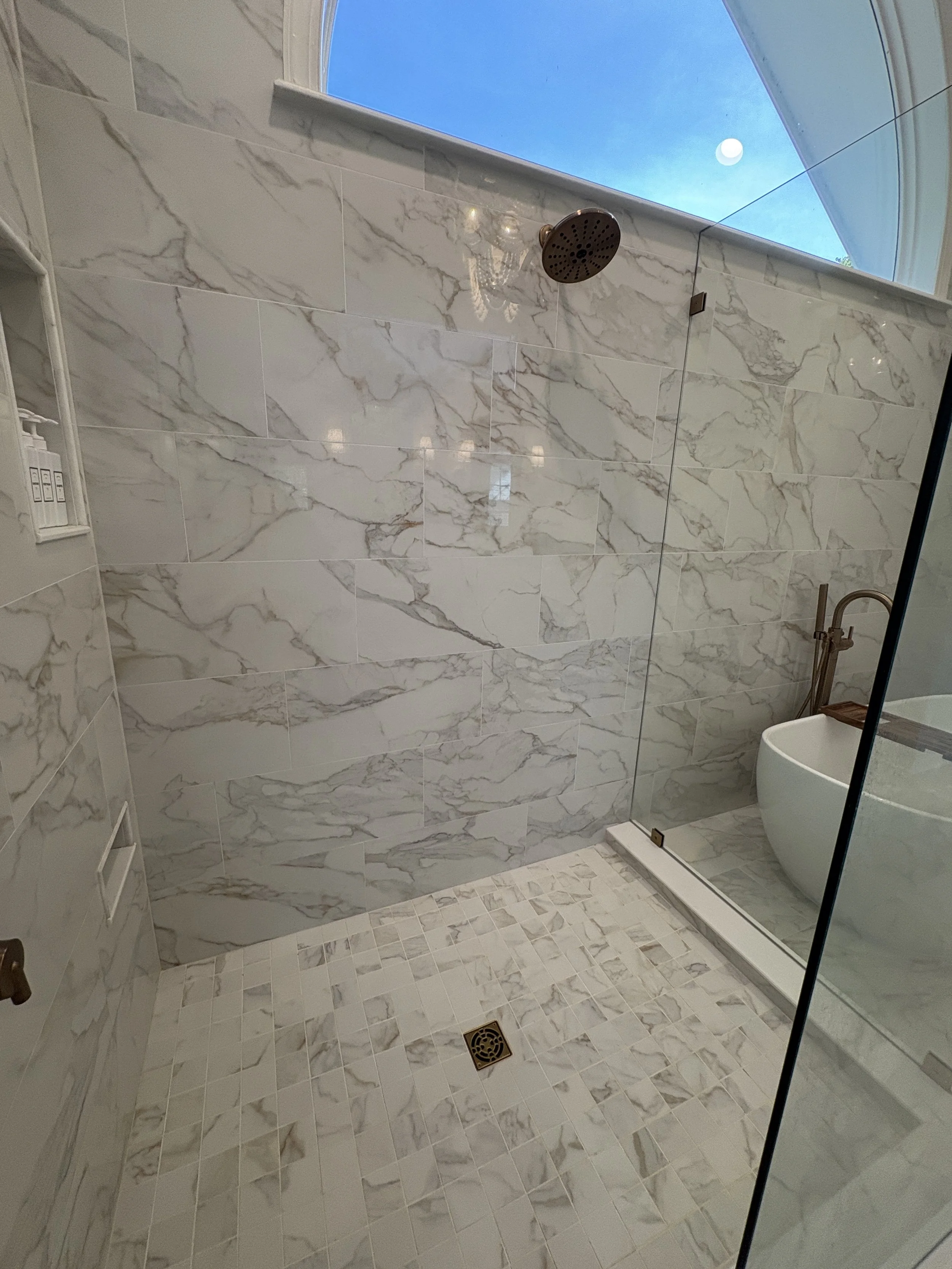 Finished custom walk-in shower featuring floor-to-ceiling marble-look porcelain tile, a frameless glass enclosure, and a seamlessly integrated tiled shower floor for a clean, modern aesthetic. Premium bronze fixtures, large-format tile installation, 