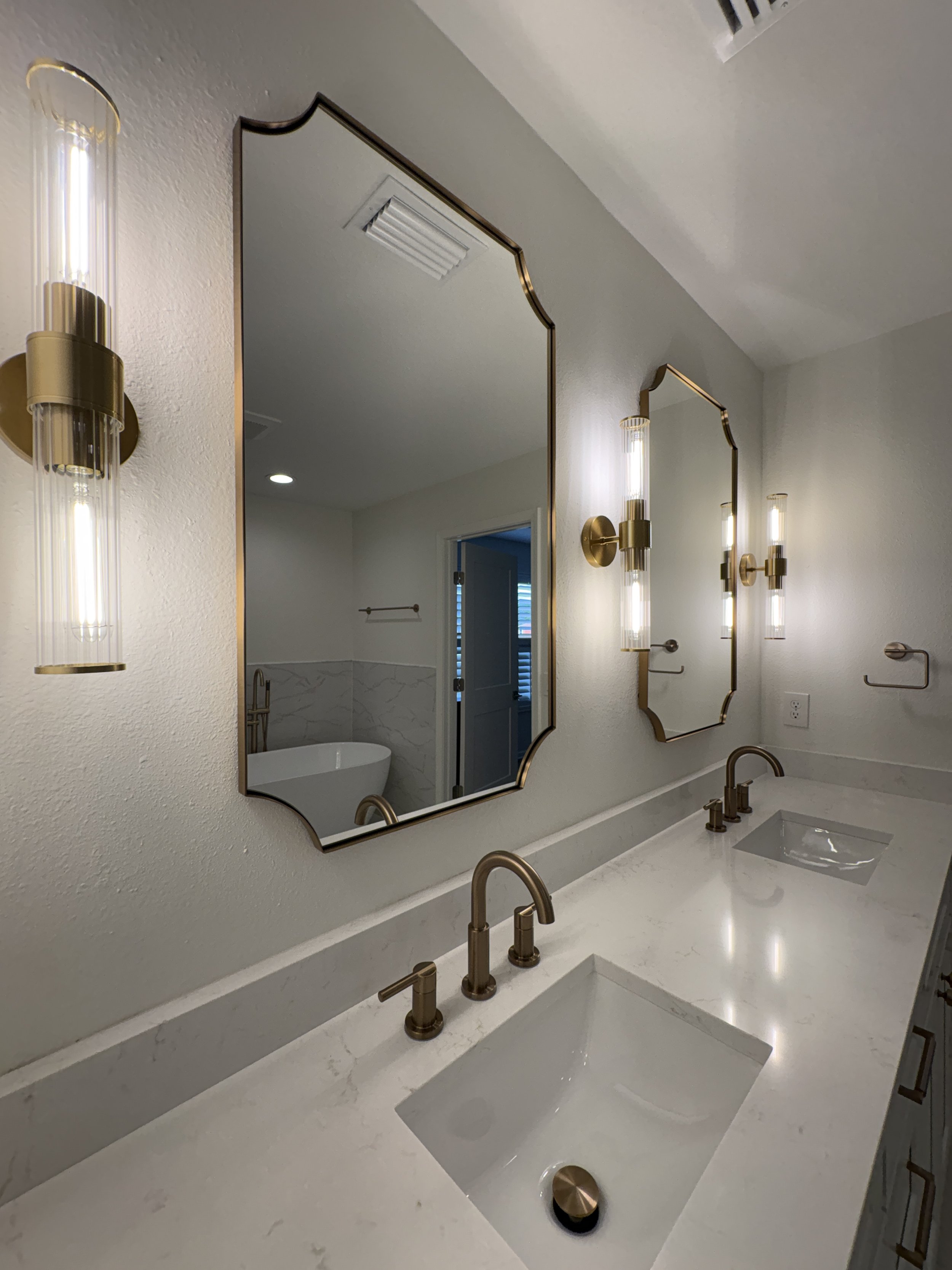 A custom double vanity with extensive storage, a sleek white countertop, decorative mirrors, and brushed gold fixtures brings elegance and function to this luxury Jacksonville condo bathroom remodel by Elite Construction of JAX.