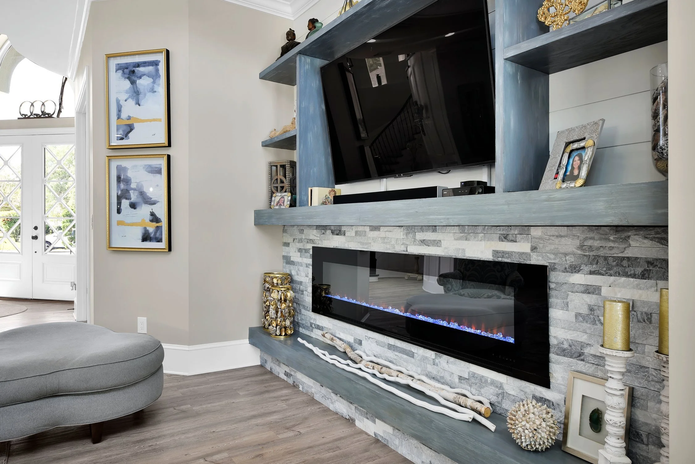 Living room with a modern fireplace built into a gray stone wall, flat-screen TV above the fireplace, blue-gray wooden shelves holding decorative items, picture frame, and framed photograph, abstract blue and gold artwork on the wall, and a gray upho