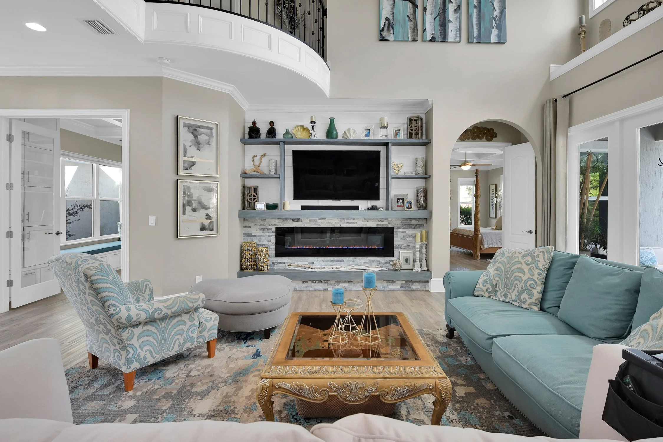 Living room with a fireplace, television above, and built-in shelves with decorations. There's a light blue sofa, a patterned armchair with a matching ottoman, a glass-topped wooden coffee table with candles, and a rug on wooden flooring. Large windo