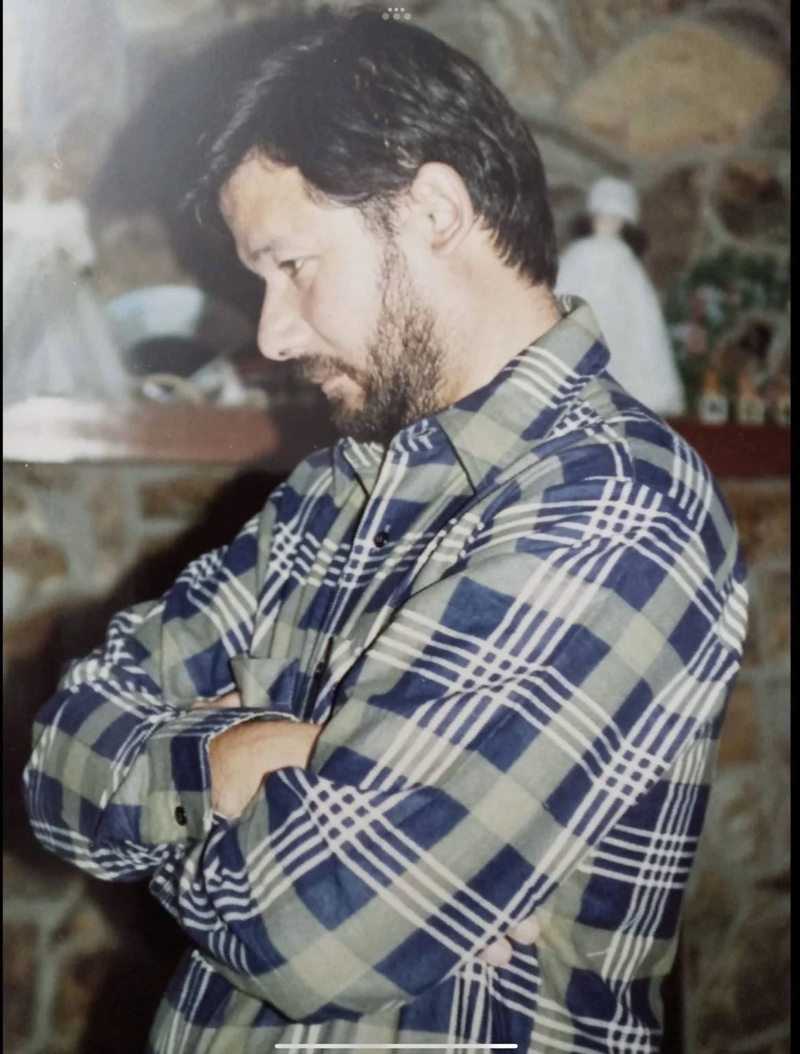 A man with dark hair and a beard, wearing a plaid shirt, standing with arms crossed in front of a stone wall, looking down.