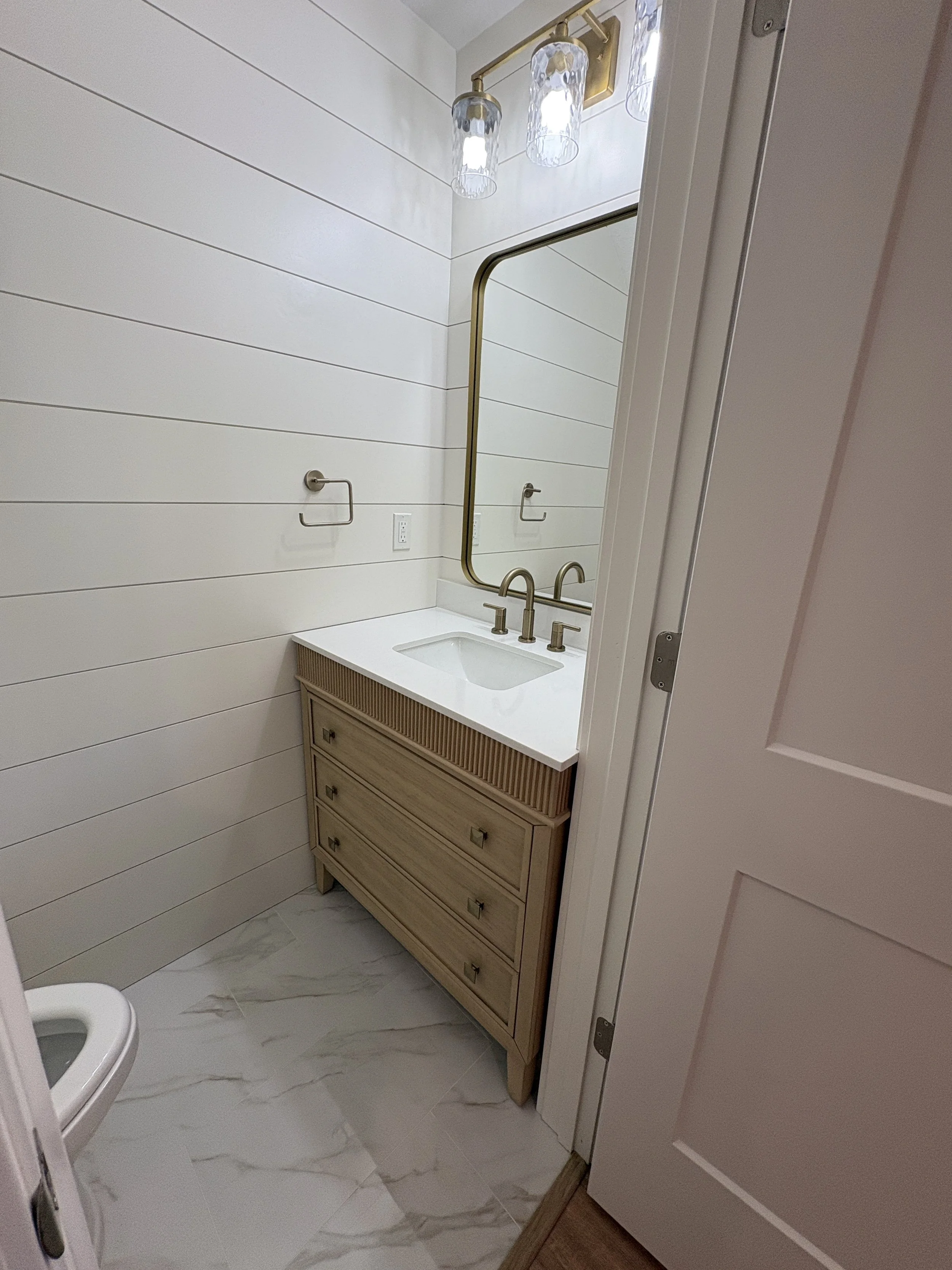 A custom wood vanity, marble-look tile flooring, shiplap walls, and modern fixtures create a warm, refined bathroom in this high-end Jacksonville condo remodel by Elite Construction of JAX.